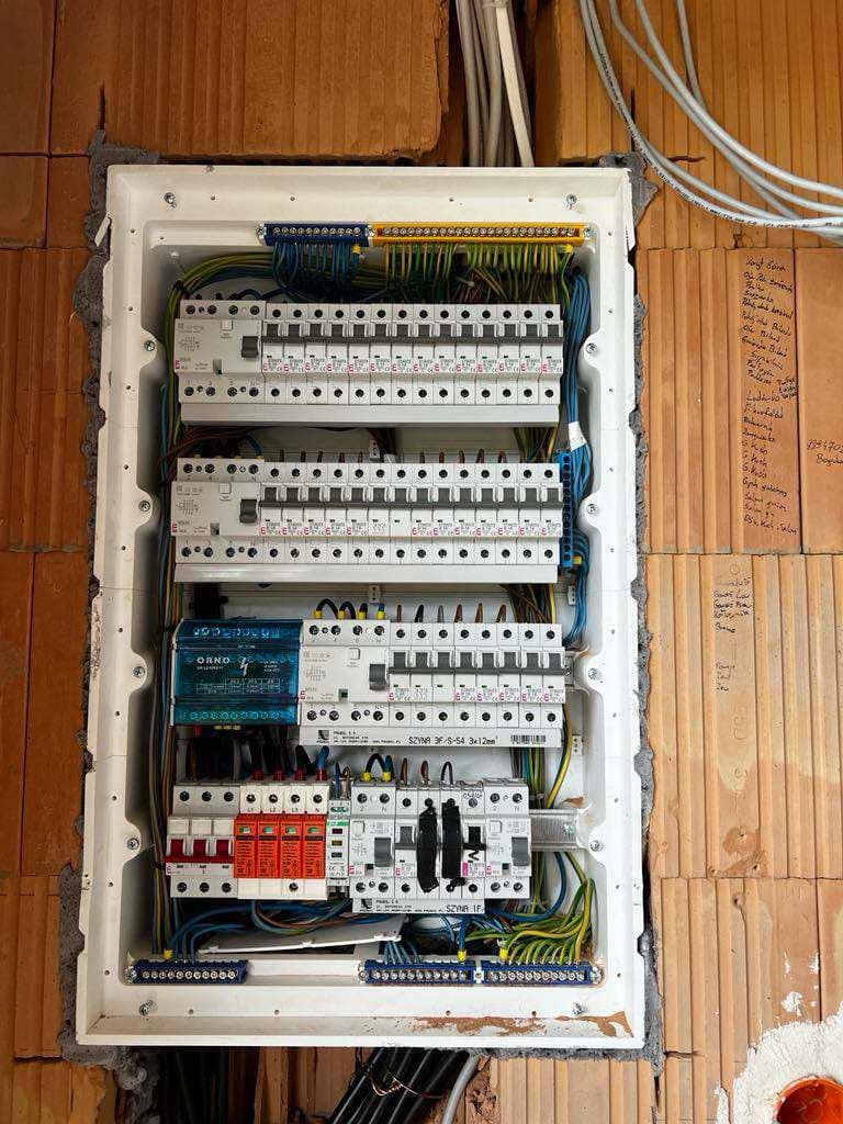 Rozdzielnica elektryczna ORNO zamontowana w surowej ścianie z cegły, widoczne liczne bezpieczniki, przewody i listwy zaciskowe.