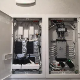 Otwarta, biała szafka rozdzielcza z widocznymi kablami, routerami TP-Link, listwą zasilającą i multiswitchem Signal MRP-512, zamontowana na szarej ścianie.