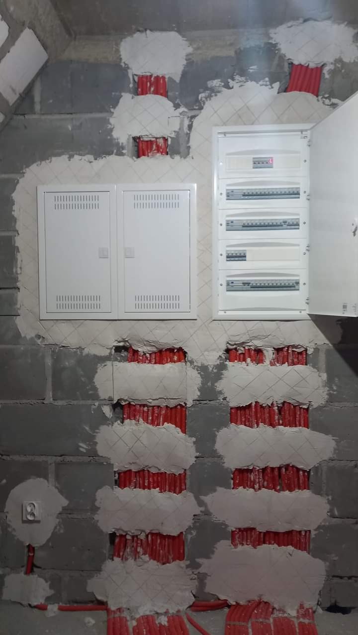 Ściana z szarego betonu komórkowego w trakcie prac, widoczne korytka z czerwonymi kablami zabezpieczone siatką i obłożone gipsem, dwie białe skrzynki elektryczne, jedna z otwartymi drzwiczkami.