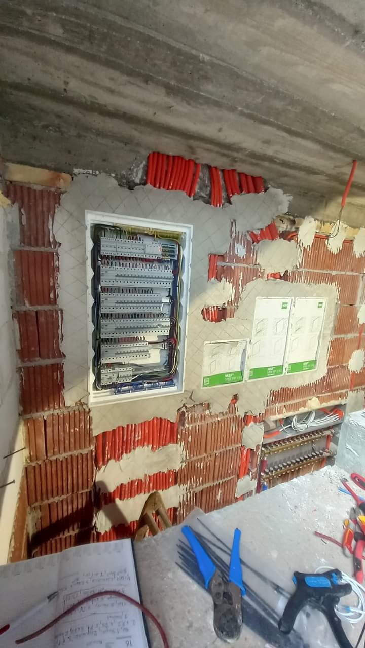 Montaż rozdzielnicy elektrycznej w surowym stanie, widoczne przewody w peszlach, częściowo otynkowana ściana z czerwonej cegły oraz notatnik i narzędzia na pierwszym planie.