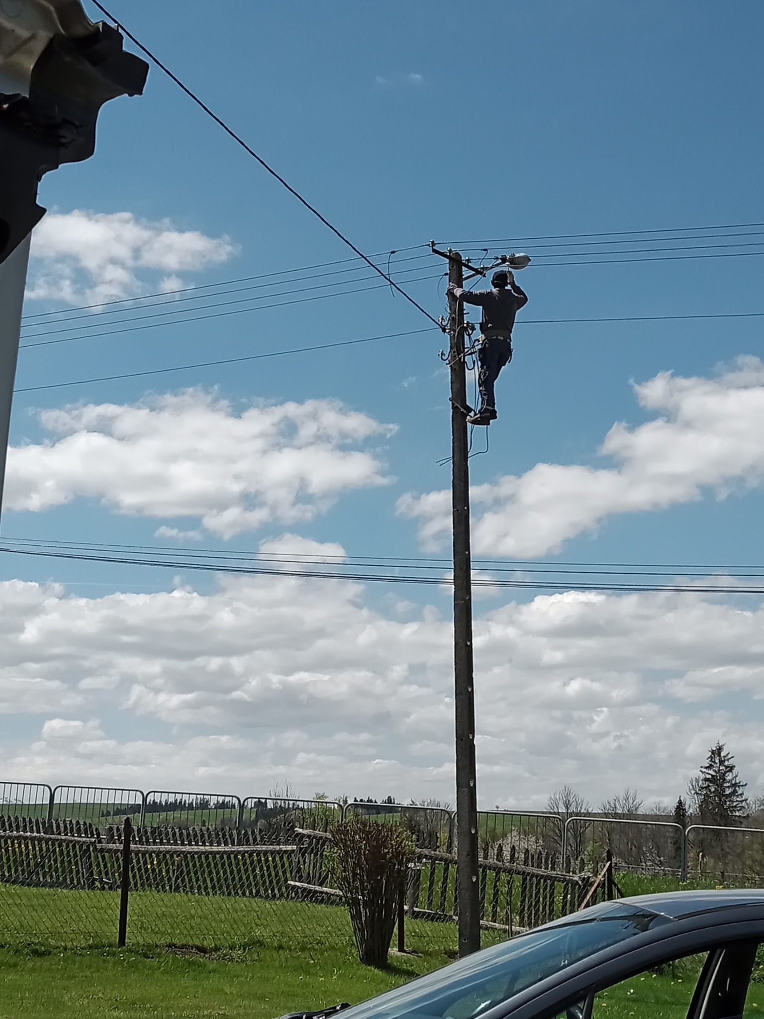 Elektryk na słupie oświetleniowym, montujący lub naprawiający lampę uliczną, na tle błękitnego nieba z chmurami. Widoczny fragment samochodu i ogrodzenie w tle.