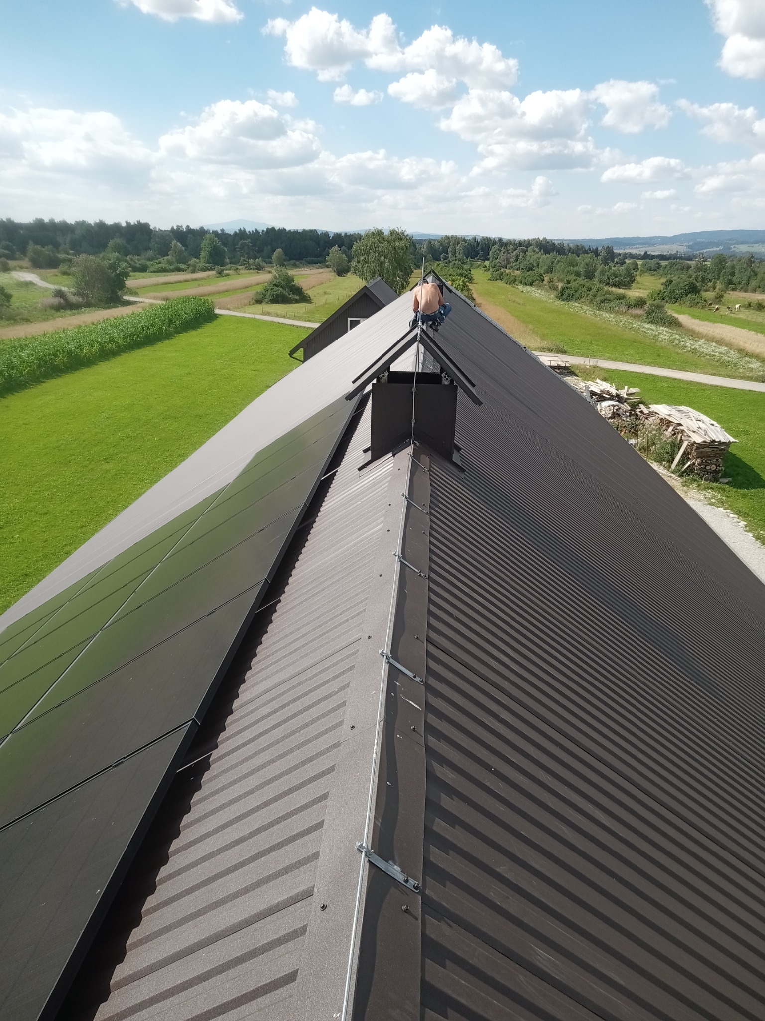 Widok z góry na dach z blachy trapezowej z zamontowanymi panelami fotowoltaicznymi i kominem; na dachu pracuje monter bez koszulki, w tle zielone pola i las.
