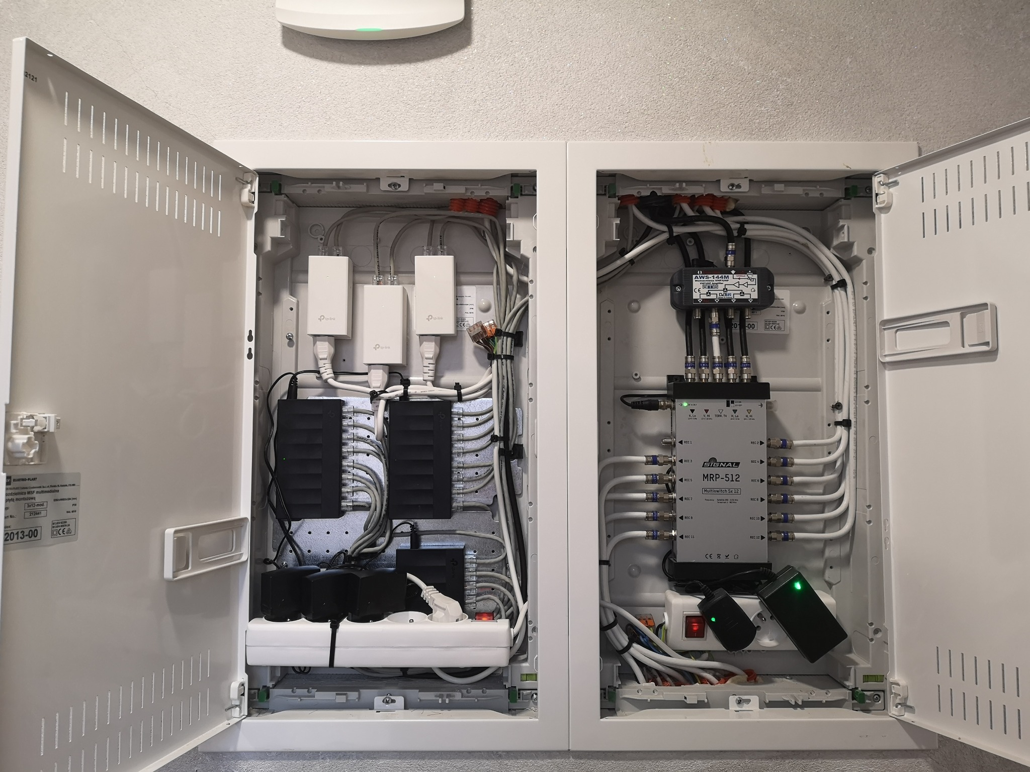 Otwarta, biała szafka rozdzielcza z widocznymi kablami, routerami TP-Link, listwą zasilającą i multiswitchem Signal MRP-512, zamontowana na szarej ścianie.