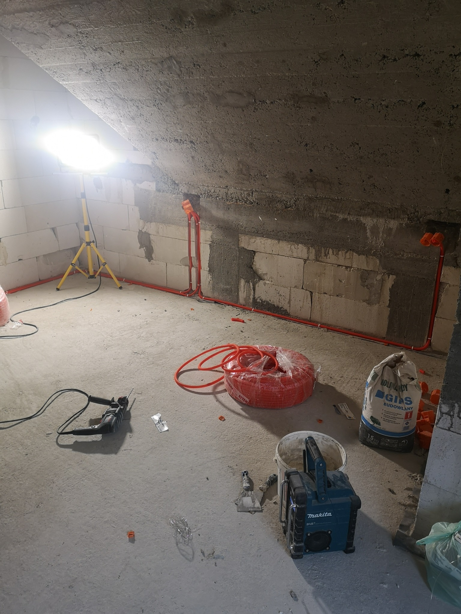 Surowy strych w trakcie prac wykończeniowych: widoczne przewody elektryczne w peszlach, wiertarka, zwój pomarańczowych kabli, worek zaprawy budowlanej oraz przenośny głośnik Makita