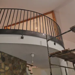 Częściowy widok metalowej balustrady na antresoli z widocznymi elementami konstrukcyjnymi i rusztowaniem malarskim, w tle ściana z kamienia i schody.