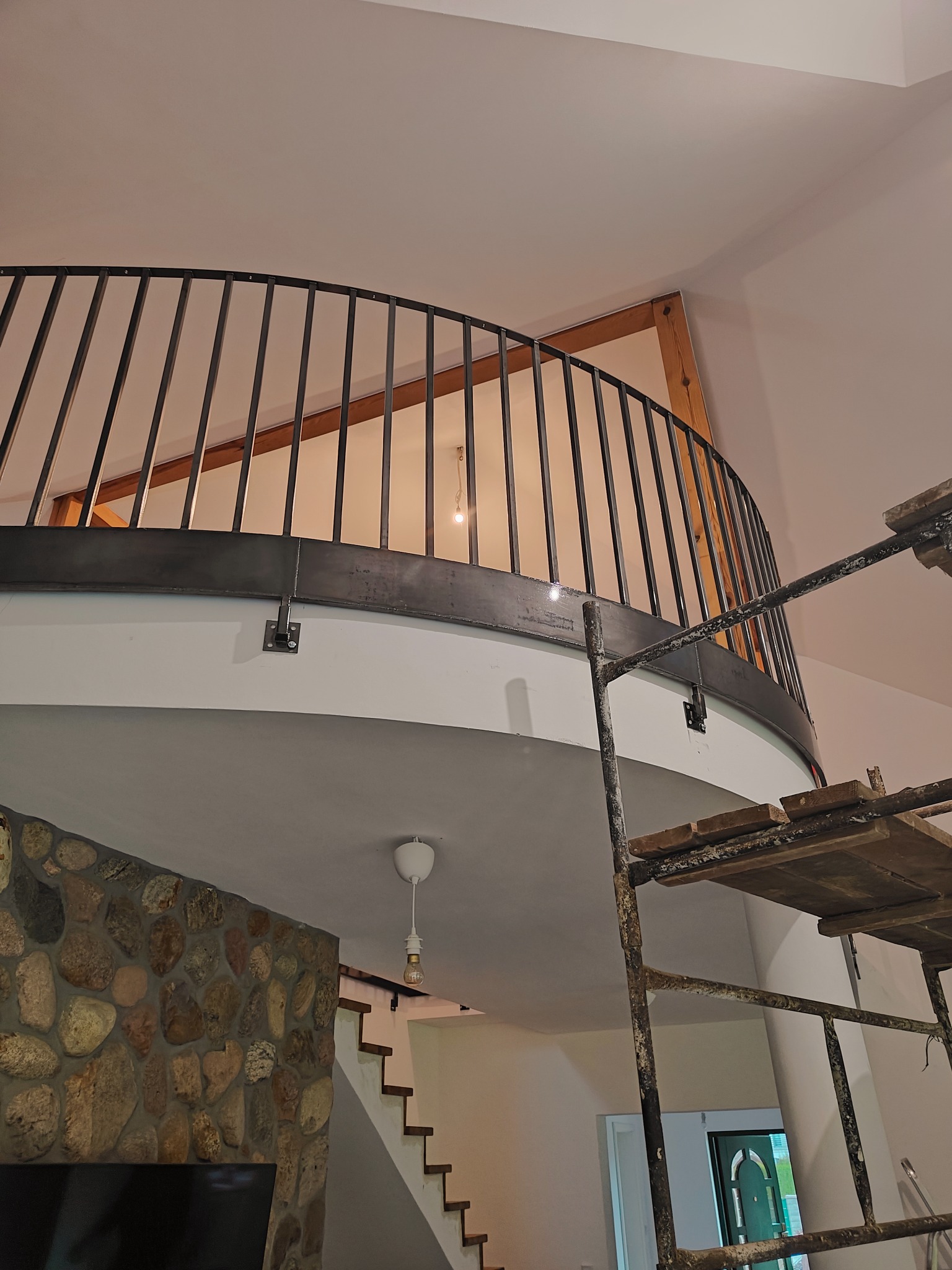 Częściowy widok metalowej balustrady na antresoli z widocznymi elementami konstrukcyjnymi i rusztowaniem malarskim, w tle ściana z kamienia i schody.