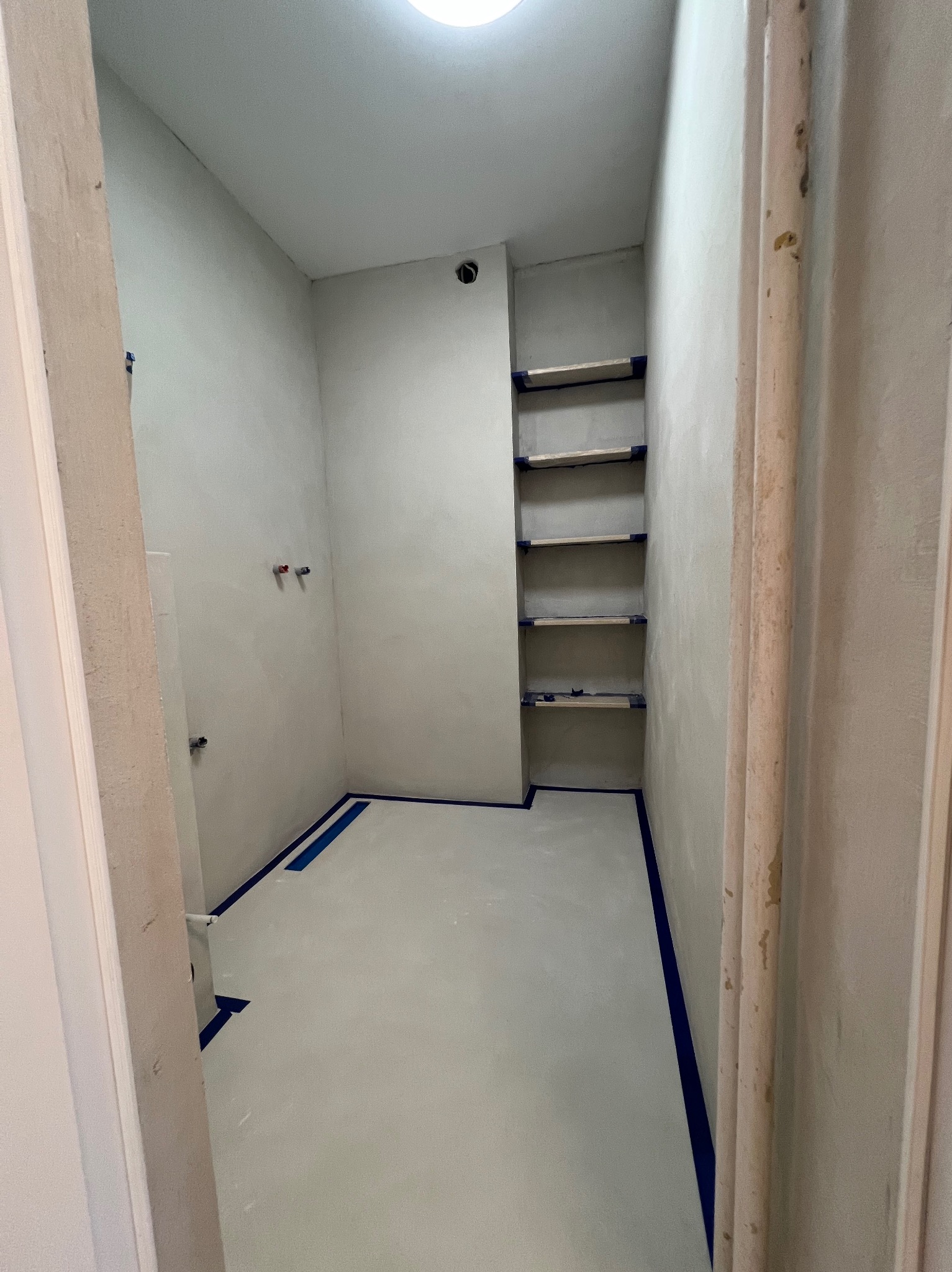 Surowe wnętrze łazienki w trakcie remontu, ściany i podłoga przygotowane pod wykończenie, zabezpieczone niebieską taśmą malarską, widoczne pionowe półki w ścianie oraz wystające rury instalacji...