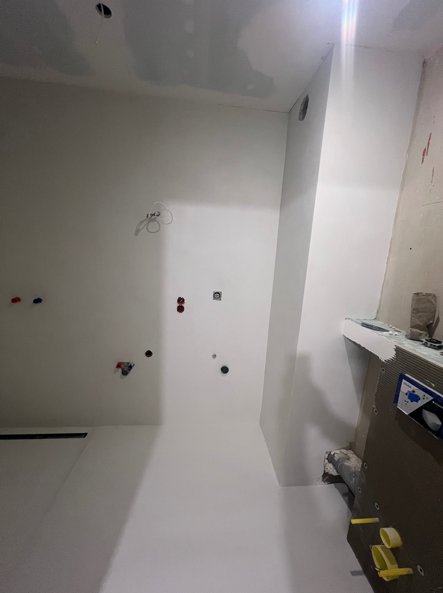 Wnętrze łazienki w trakcie remontu, widoczne przygotowane punkty hydrauliczne i elektryczne na ścianie, świeżo pomalowane ściany i sufit, oraz fragment zabudowy stelaża podtynkowego WC z elementami...