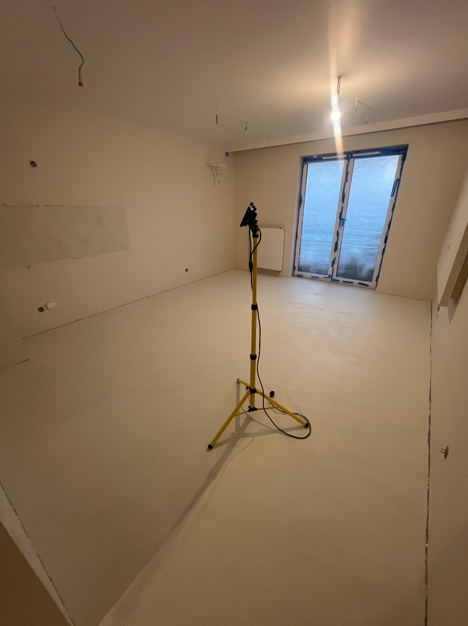 Wnętrze pokoju z nową jasną posadzką, zabezpieczone okno folią, widoczna lampa budowlana na trójnogu i niedokończone ściany.