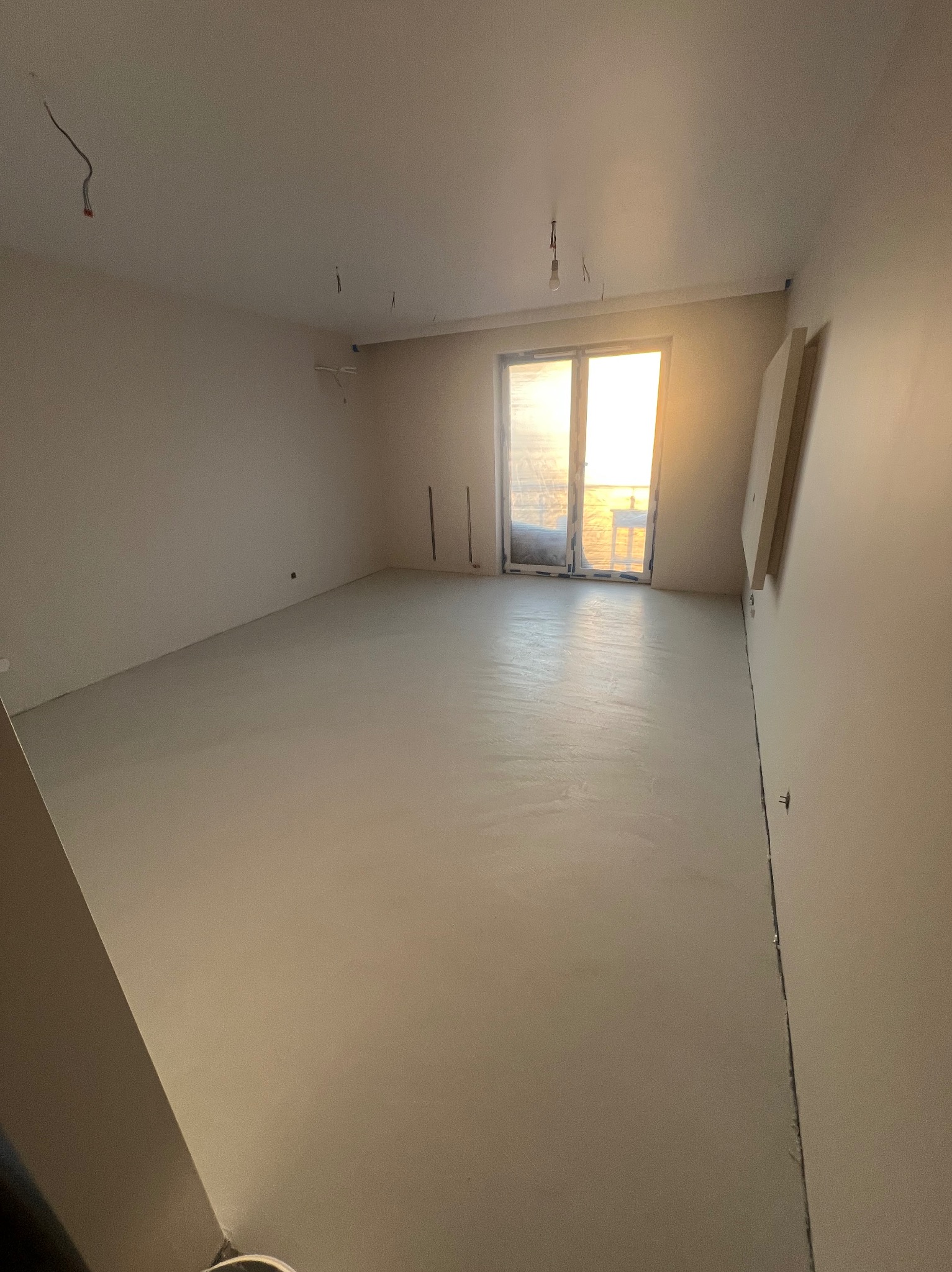 Jasne wnętrze pokoju po wylaniu posadzki samopoziomującej, widoczne niedokończone prace elektryczne przy suficie i surowe ściany przygotowane do malowania.