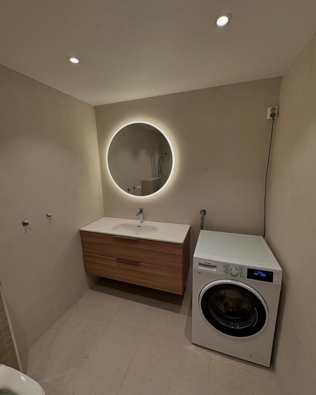 Nowoczesna łazienka z umywalką na szafce z drewnianymi lamelami, okrągłym lustrem z podświetleniem LED i białą pralką.