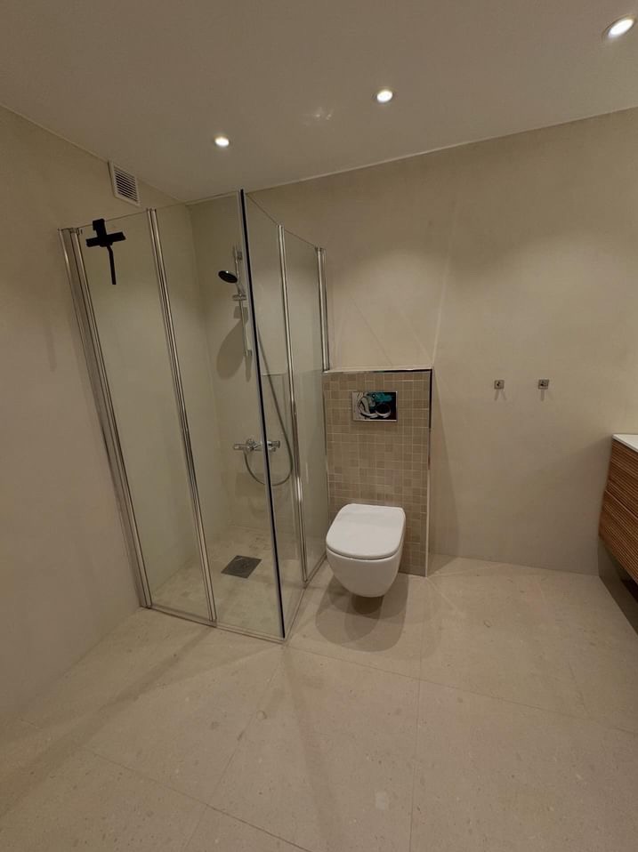 Nowoczesna łazienka z kabiną prysznicową ze szkła i metalu, wiszącą toaletą obudowaną mozaiką i beżową podłogą z delikatnym wzorem.