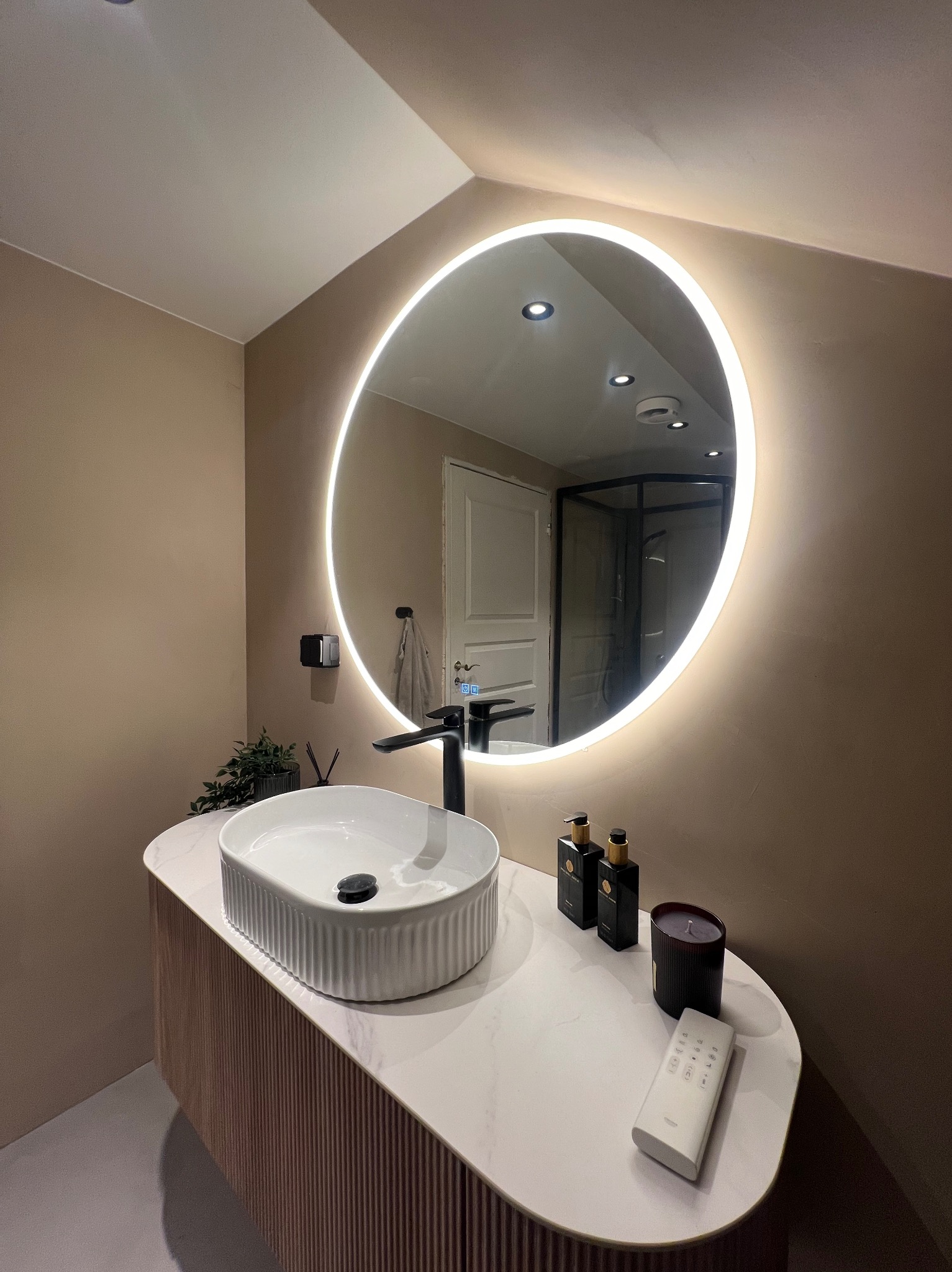 Nowoczesna łazienka z okrągłym lustrem podświetlanym LED, białą umywalką nablatową z czarną armaturą, blatem imitującym marmur, beżowymi ścianami i widoczną kabiną prysznicową w tle.