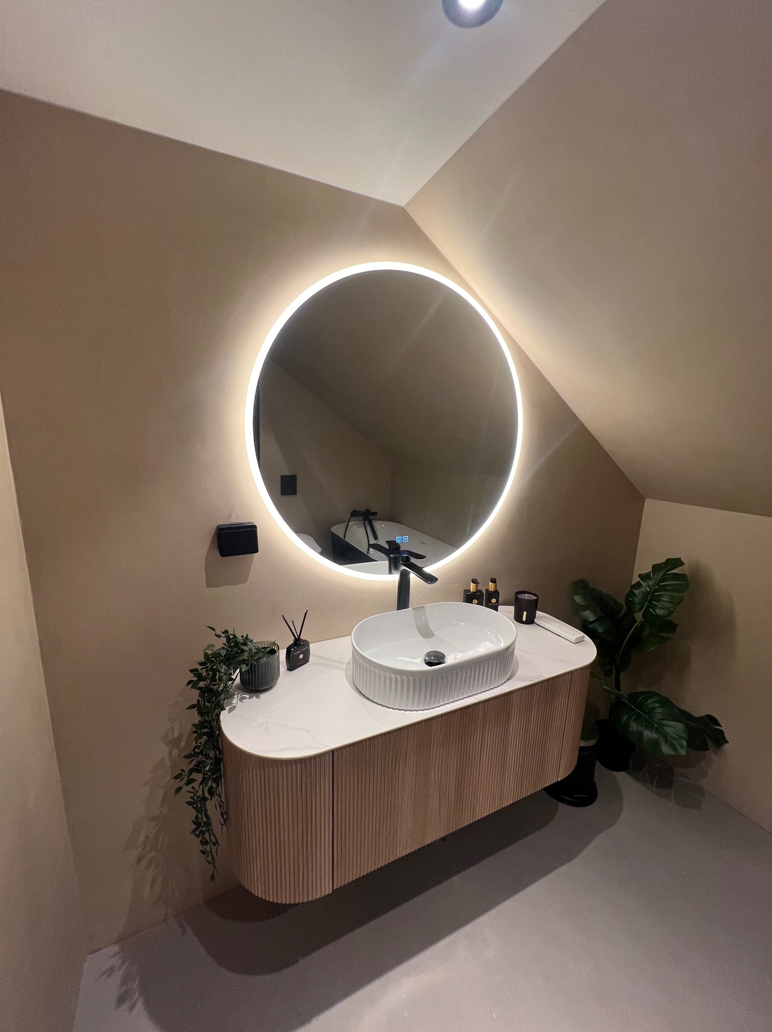 Nowoczesna łazienka na poddaszu ze skośnym sufitem, umywalką nablatową o żłobkowanej strukturze, szafką z ryflowanymi frontami, okrągłym lustrem z podświetleniem LED, roślinami doniczkowymi...