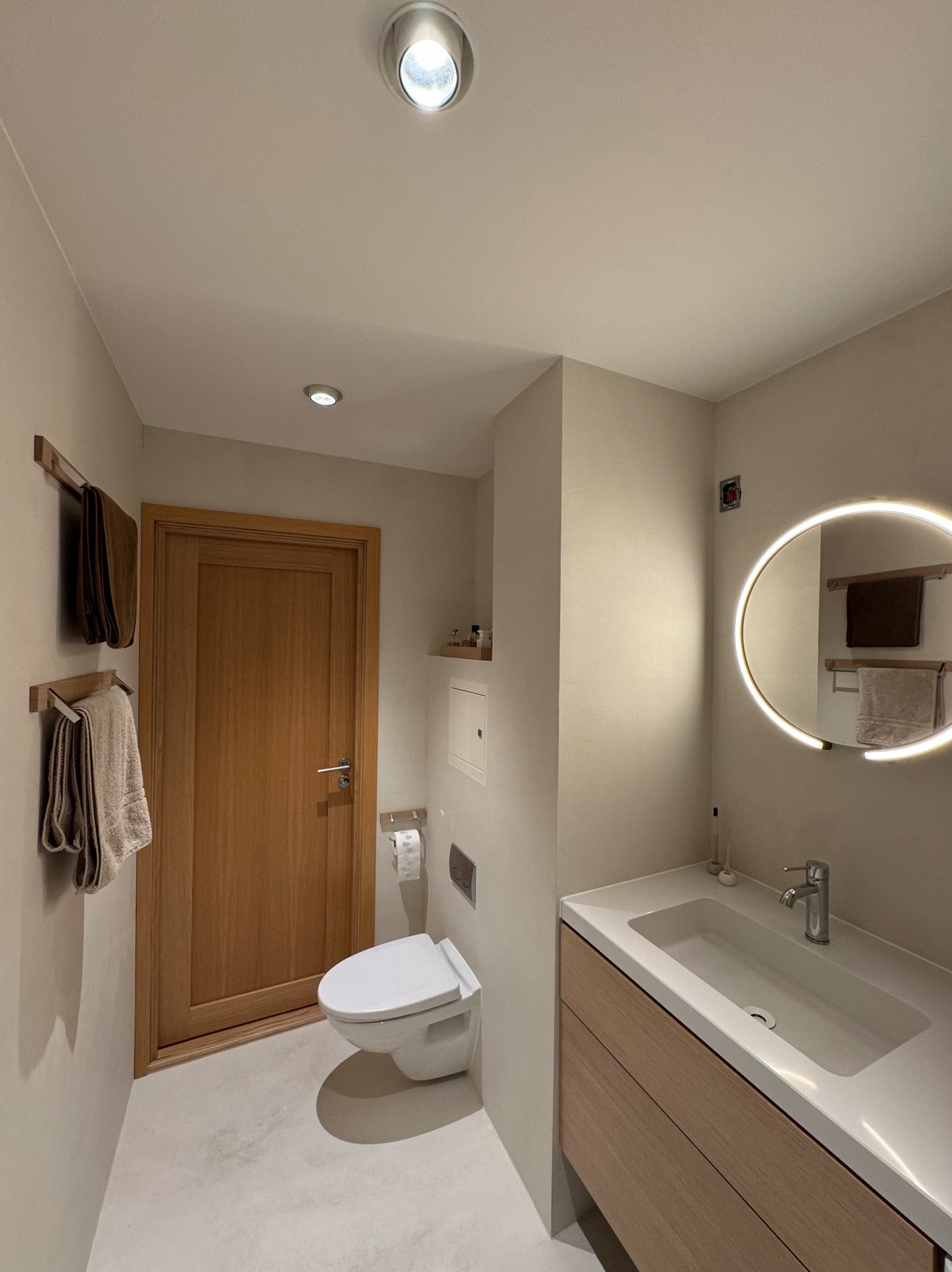 Nowoczesna łazienka z beżowymi ścianami, podwieszaną toaletą, drewnianą szafką pod umywalką i okrągłym lustrem z podświetleniem LED. Widoczne drewniane wieszaki na ręczniki.