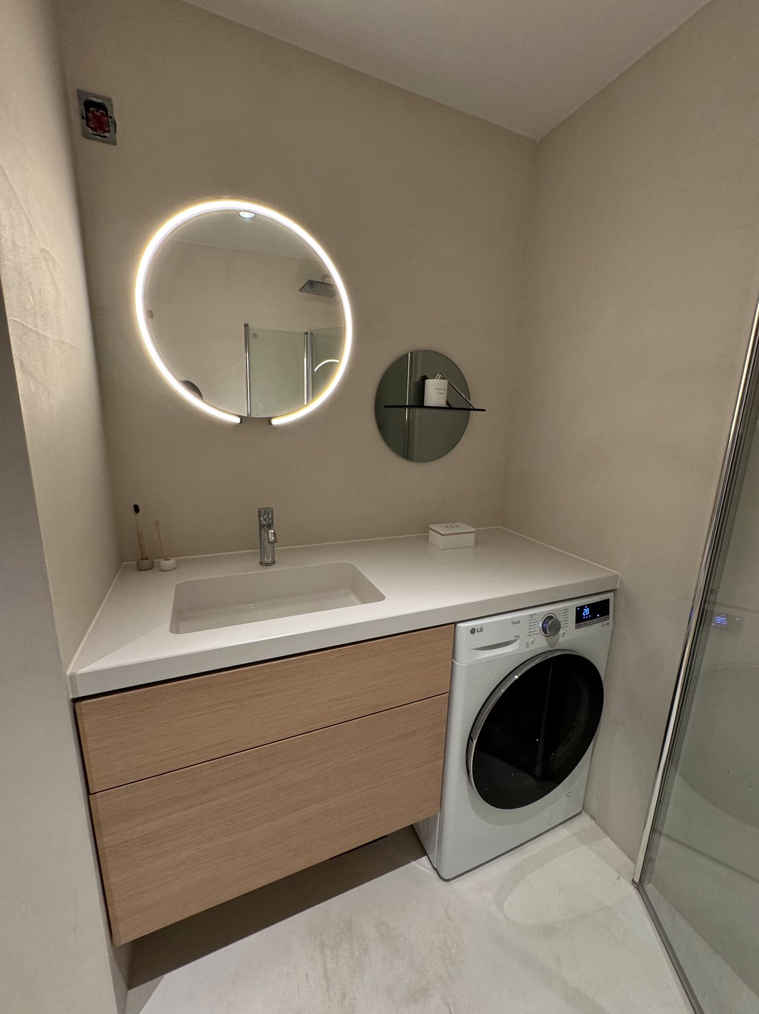 Nowoczesna łazienka z umywalką wpuszczoną w blat, szafką z drewnopodobnymi frontami, okrągłym lustrem z podświetleniem LED i pralką LG obok kabiny prysznicowej.