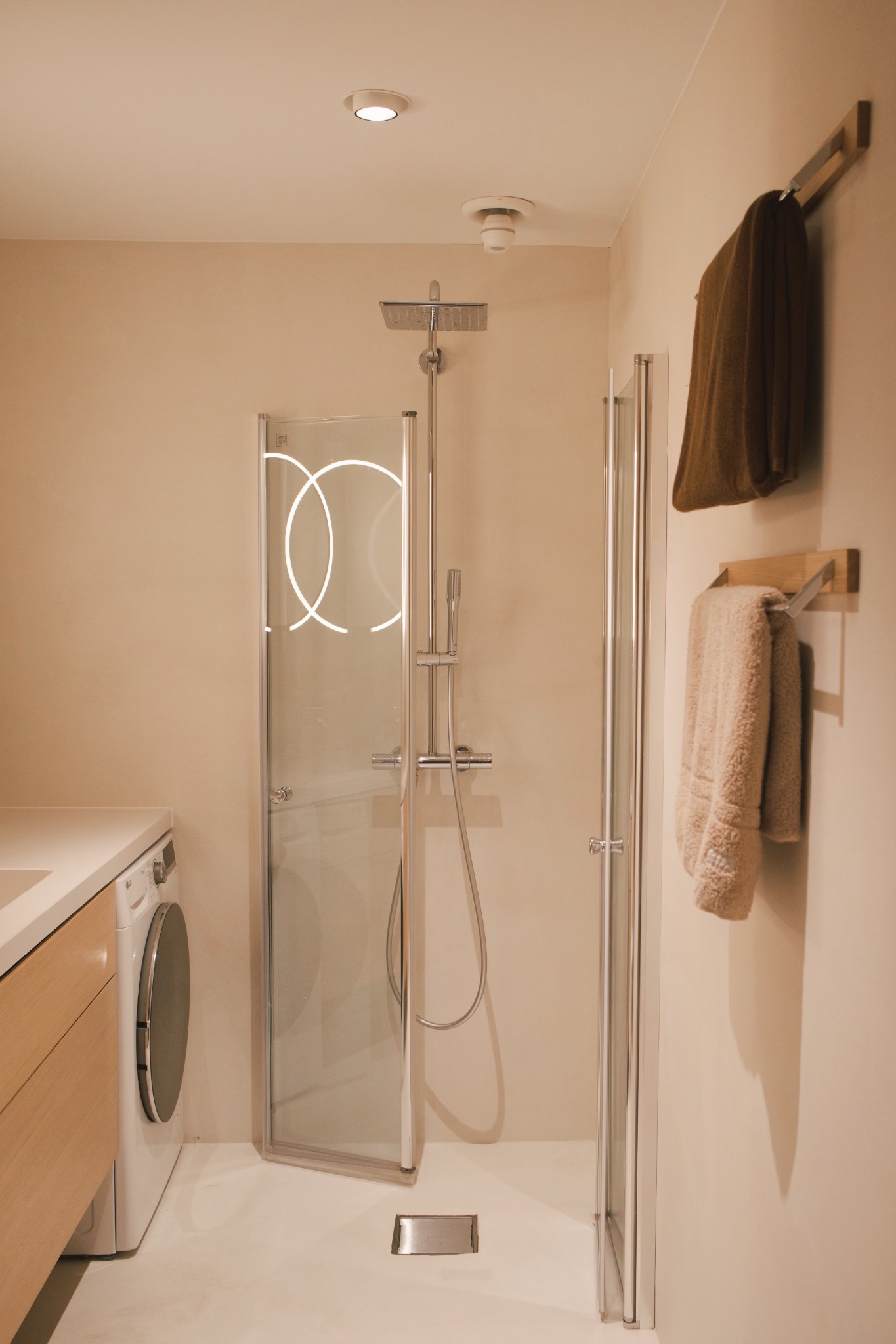 Nowoczesna łazienka z kabiną prysznicową z podświetlanymi elementami, pralką pod blatem i ręcznikami na drewnianych wieszakach.