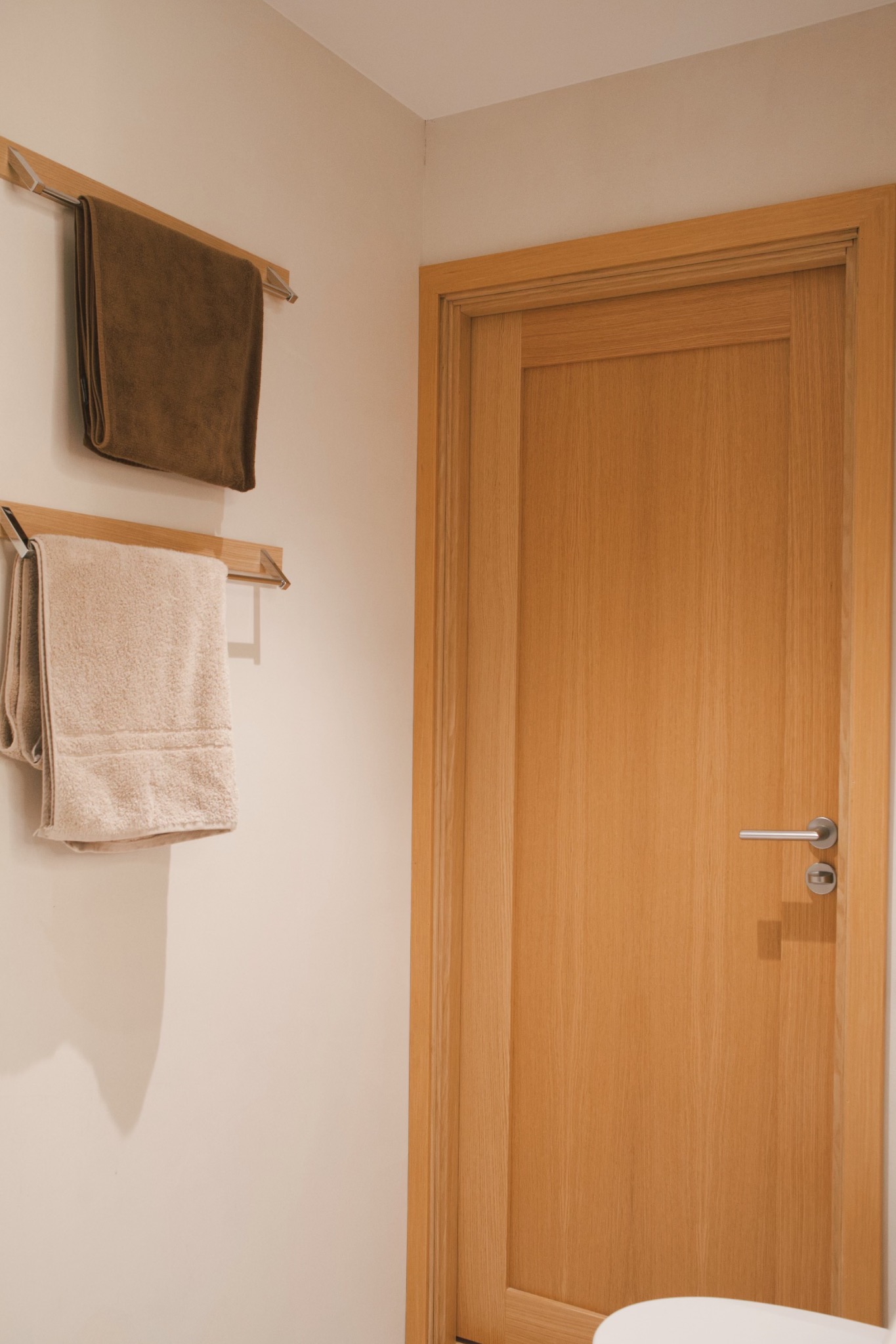 Nowoczesna łazienka z drewnianymi drzwiami, chromowanym uchwytem i dwoma ręcznikami na wieszakach: brązowym i beżowym.