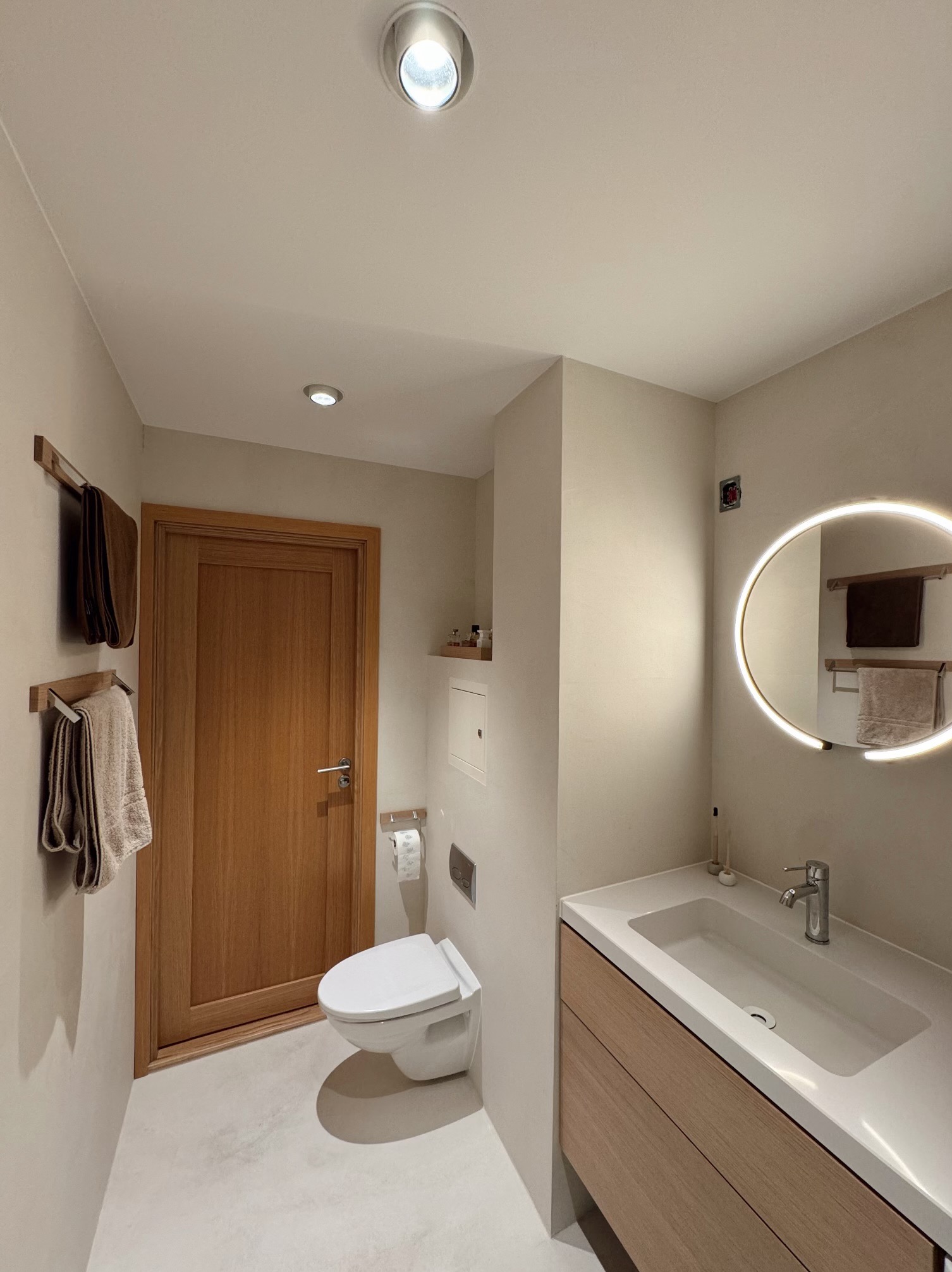 Nowoczesna łazienka z wykończeniem z mikrocementu: wisząca toaleta, umywalka z drewnianą szafką, okrągłe lustro z podświetleniem LED i drewniane wieszaki na ręczniki.