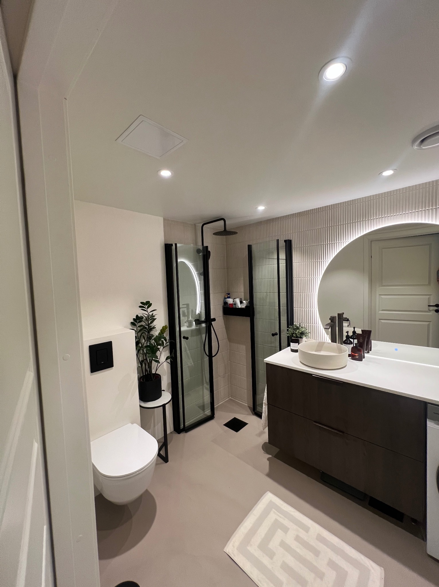 Nowoczesna łazienka w jasnych kolorach z dwoma kabinami prysznicowymi z czarnymi ramami, umywalką nablatową na szafce z ciemnego drewna, podświetlanym okrągłym lustrem i wiszącą toaletą.