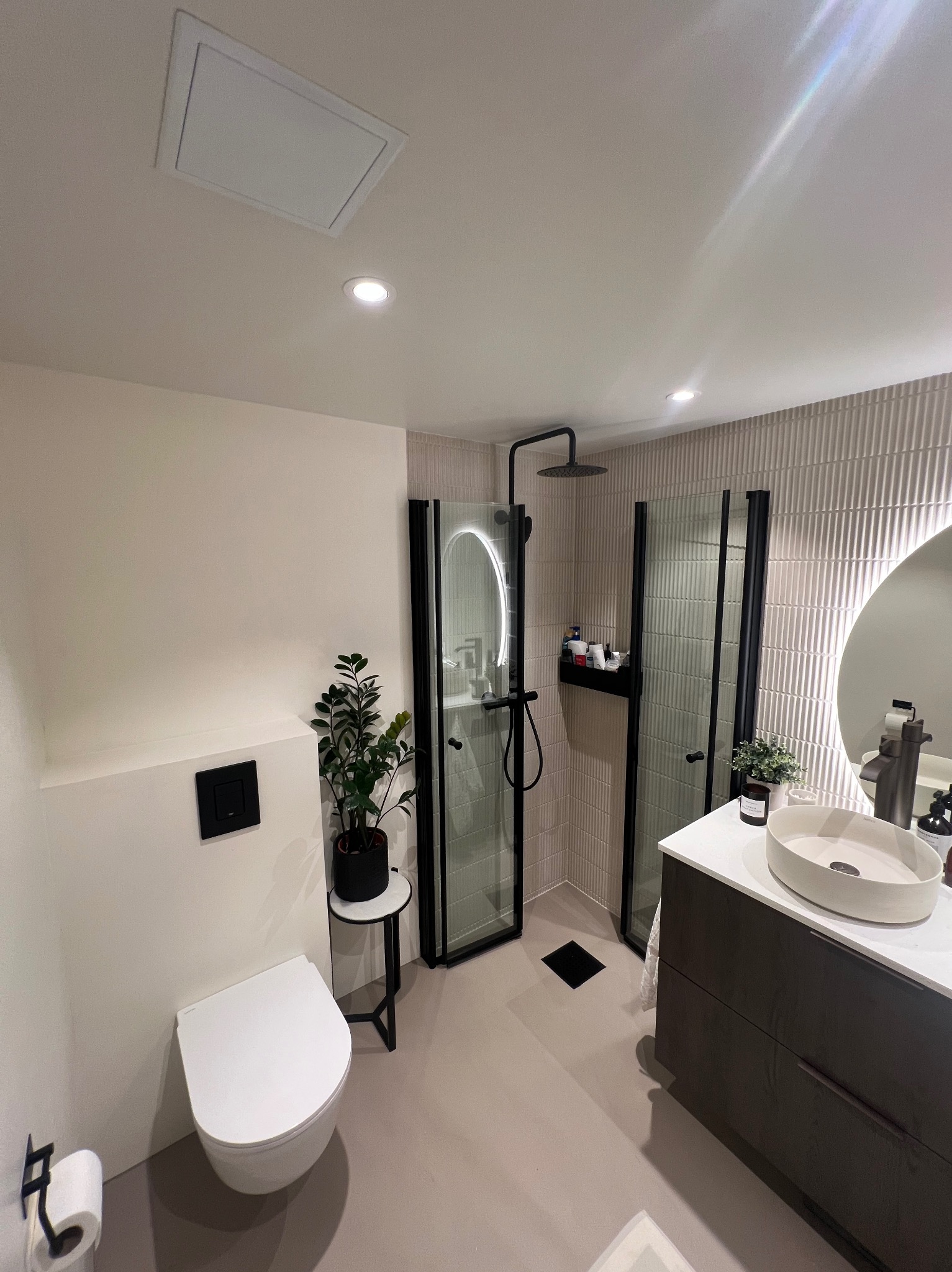 Nowoczesna łazienka z kabiną prysznicową typu walk-in z czarnymi ramami, wiszącą toaletą z czarnym przyciskiem spłukiwania, umywalką nablatową na szafce w kolorze ciemnego drewna i roślinami...