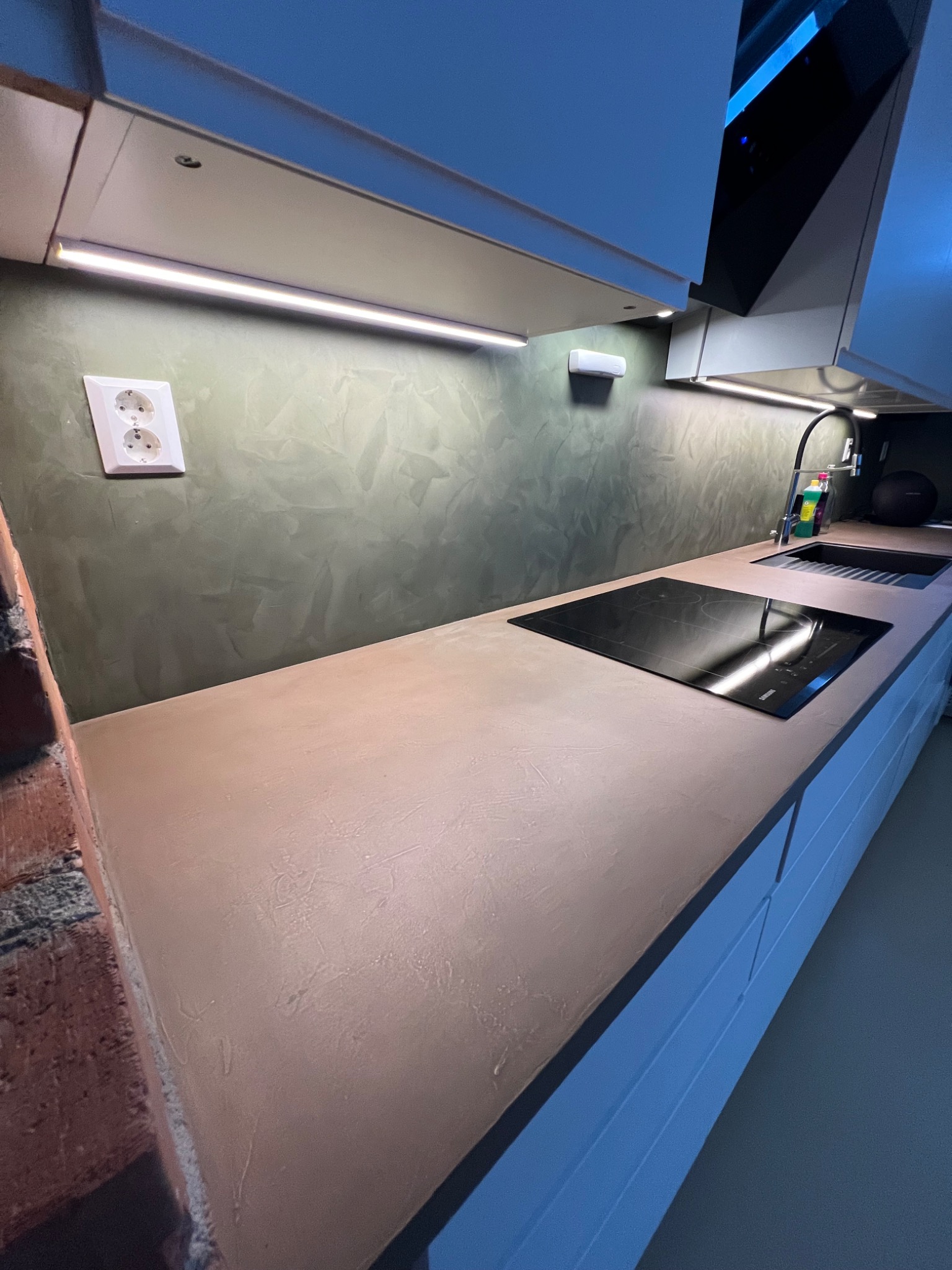 Kuchnia z blatem i ścianą wykończoną w mikrocemencie w odcieniach beżu i zieleni, widoczna płyta indukcyjna i zlew, oświetlenie LED pod szafkami.