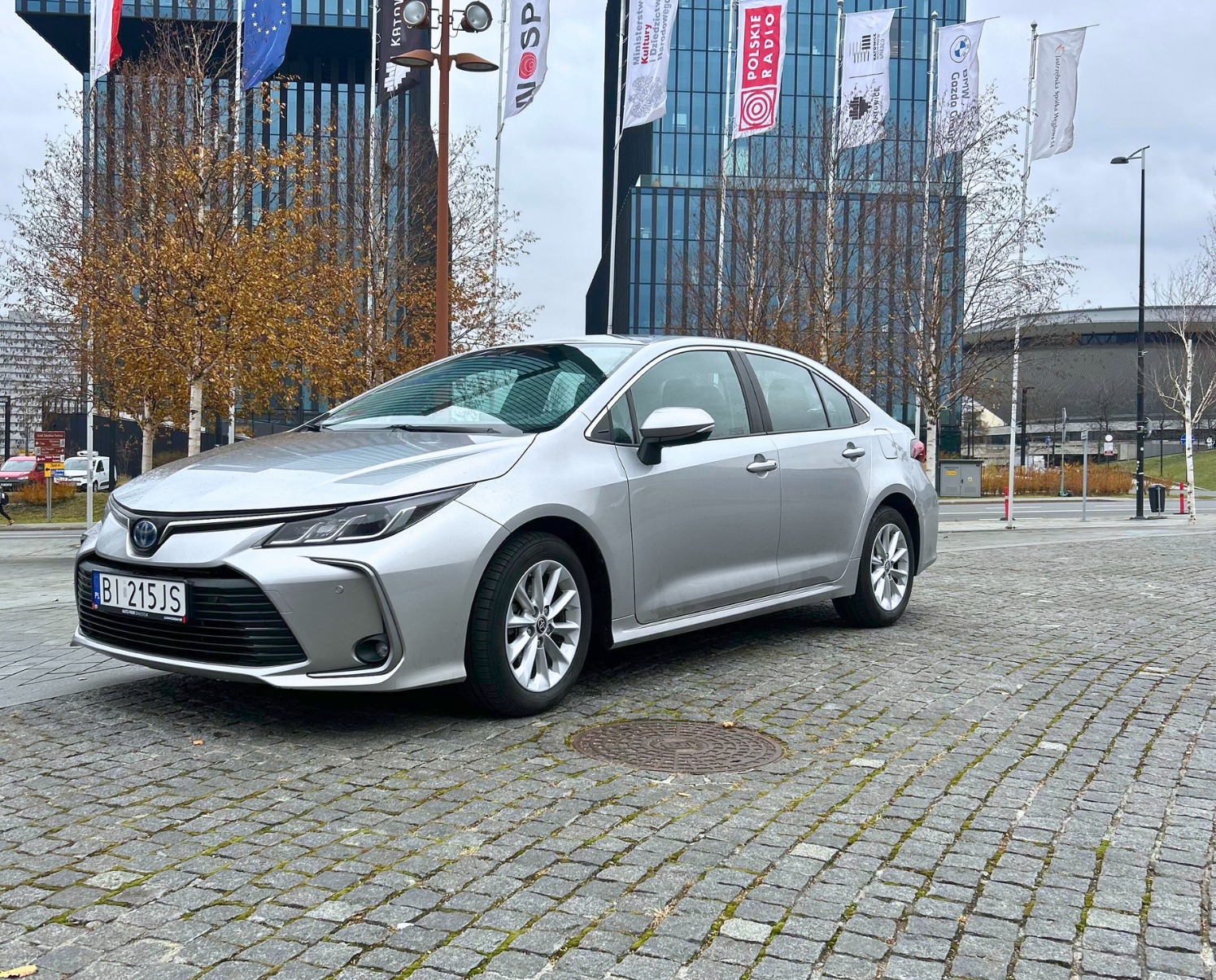 Srebrny sedan na brukowanym placu miejskim z budynkami i flagami w tle, widoczna tablica rejestracyjna BI 215JS.