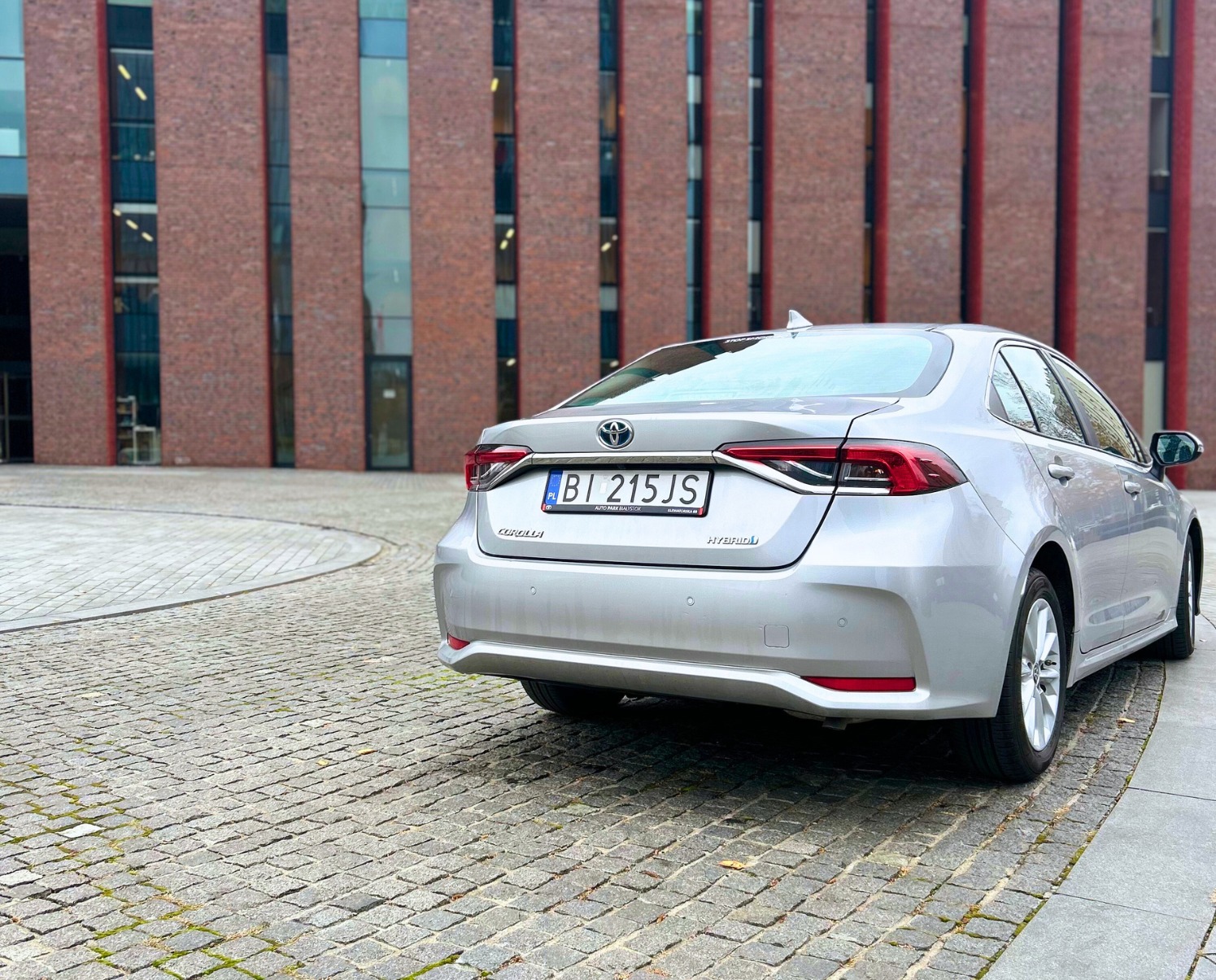 Srebrna Toyota Corolla Hybrid zaparkowana na kostce brukowej przed nowoczesnym budynkiem z cegły, widok z tyłu.
