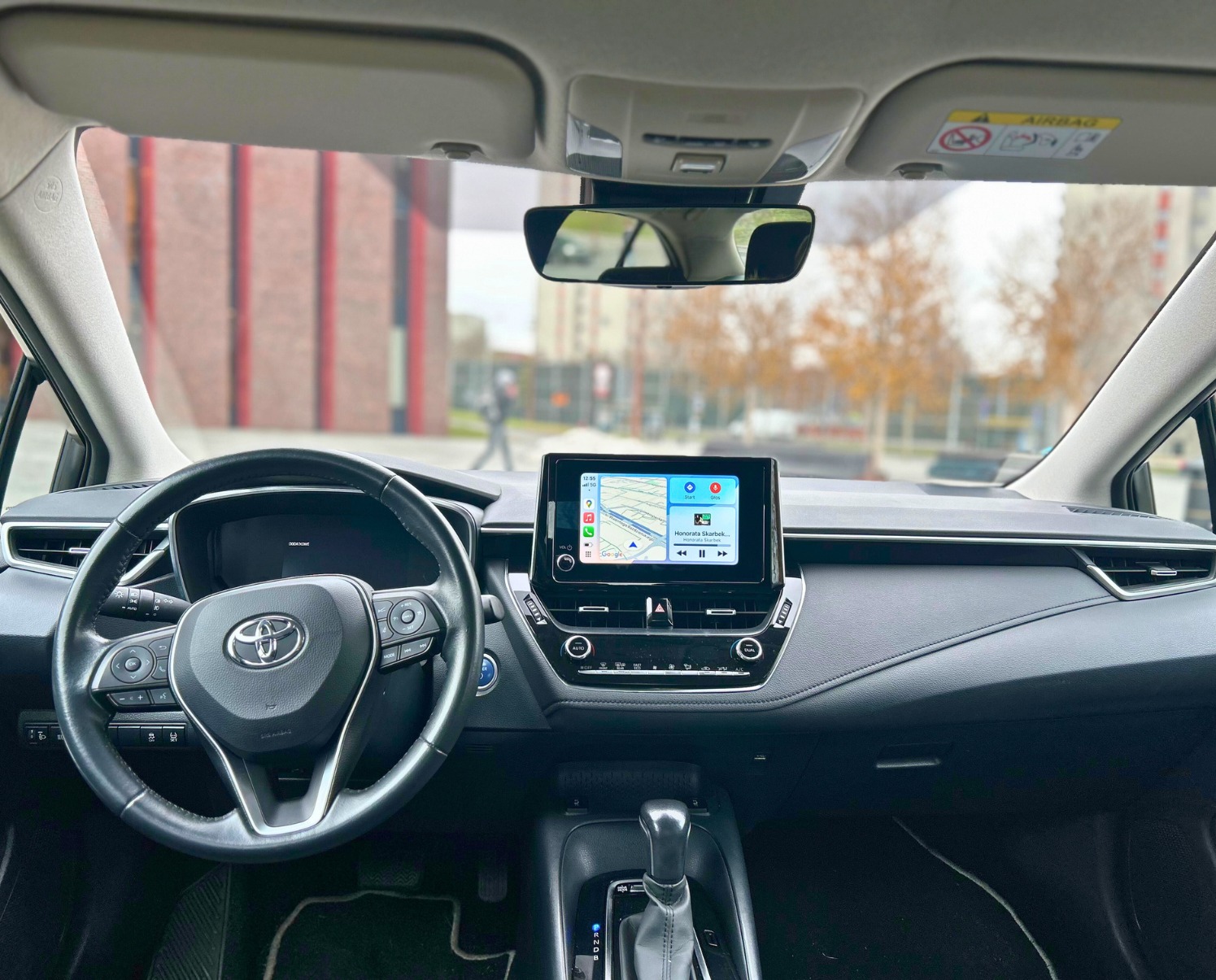 Wnętrze Toyoty Corolla z włączoną nawigacją GPS na ekranie dotykowym; widok z wnętrza pojazdu na budynek z czerwonej cegły i drzewa bez liści.