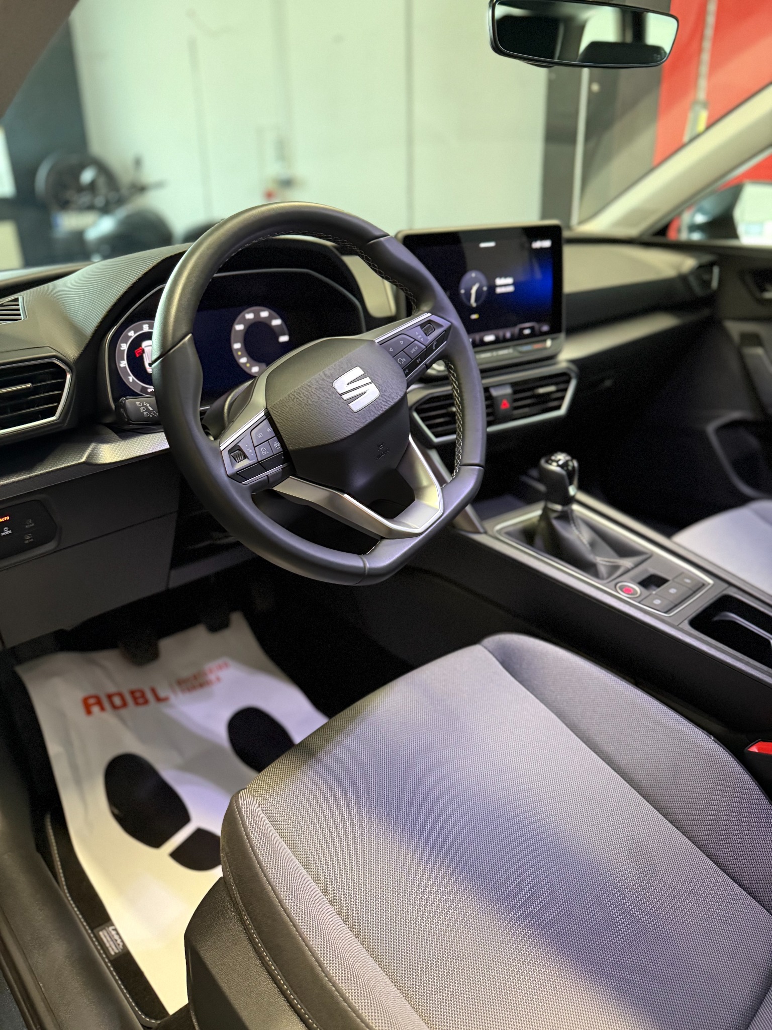 Wnętrze samochodu z kierownicą, deską rozdzielczą i siedzeniem. Na podłodze mata ochronna. Widoczny panel sterowania i dźwignia zmiany biegów.