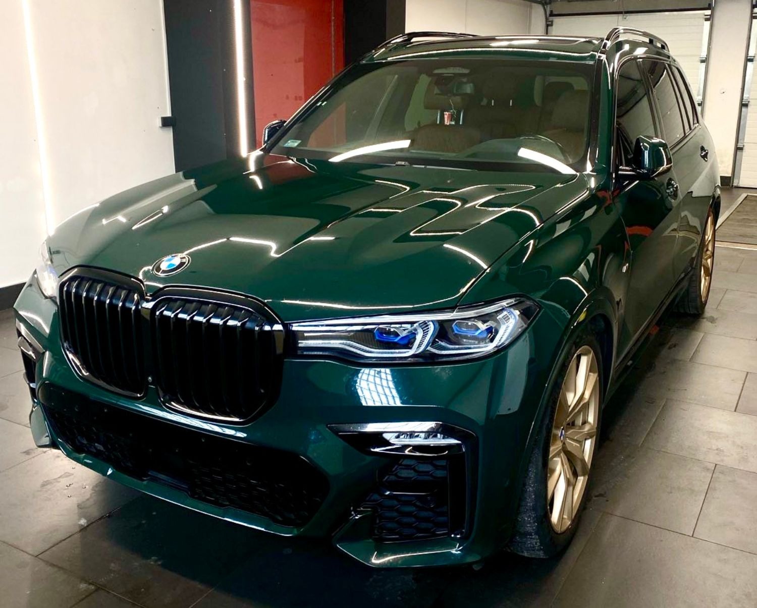 Błyszczący, ciemnozielony SUV BMW X7 po detailingu z złotymi felgami, widoczny w myjni z oświetleniem LED, z bliska, z niskiego kąta.