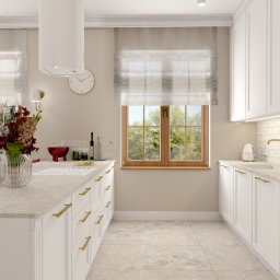 Karolina Szczesna Wnętrza - Jasna kuchnia w stylu klasycznym z białymi szafkami, złotymi uchwytami i marmurowym blatem, centralnie umieszczony wysoki bukiet kwiatów w szklanym wazonie, okno z widokiem na zieleń.