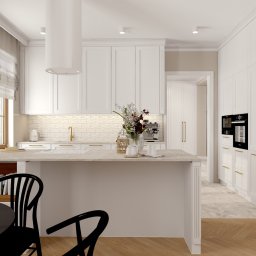 Karolina Szczesna Wnętrza - Jasna kuchnia w stylu klasycznym z wyspą, białymi szafkami, złotymi detalami i czarnymi krzesłami przy okrągłym stole. Na blacie kwiaty w wazonie.