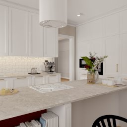 Karolina Szczesna Wnętrza - Jasna kuchnia w stylu klasycznym z wyspą, białymi szafkami z frezowanymi frontami i złotymi uchwytami, oświetleniem podszafkowym i płytkami imitującymi cegłę. Na blacie wyspy widoczna płyta...