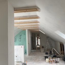 Emil Czesak - Poddasze w trakcie prac wykończeniowych: szpachlowanie ścian, drewniane belki zabezpieczone folią, drabina, narzędzia malarskie i elektryczne, widoczny pracownik na podwyższeniu.