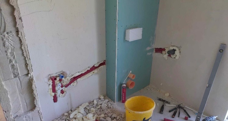 Instalacja wodno-kanalizacyjna w łazience w trakcie prac:  rury w izolacji piankowej, płyta gipsowo-kartonowa, poziomica, żółte wiadro i narzędzia na podłodze.