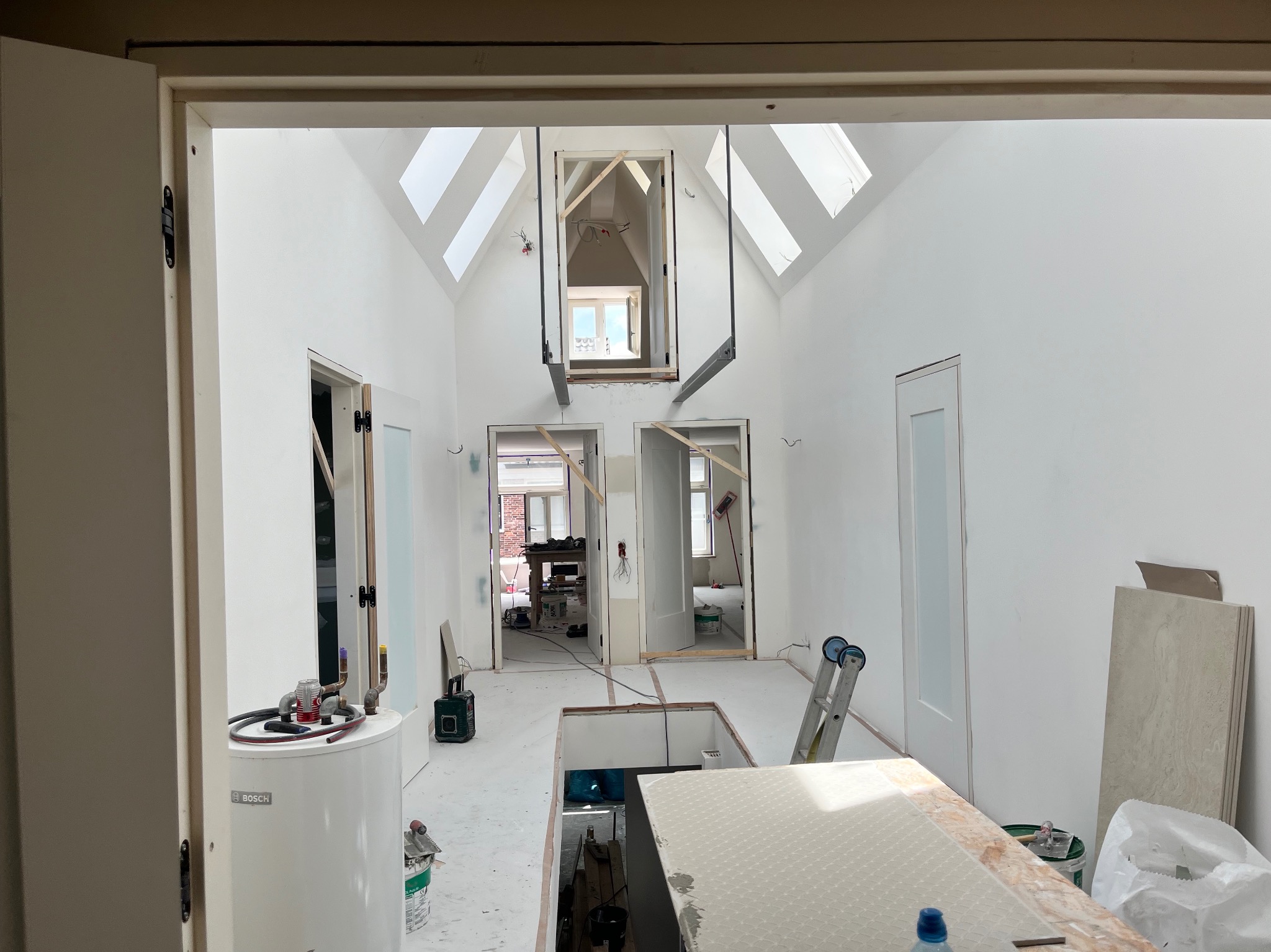 Wnętrze domu w trakcie prac wykończeniowych: białe ściany, okna dachowe, widoczne instalacje, drabina, boiler Bosch, płyty podłogowe i narzędzia, perspektywa z otwartego przejścia.