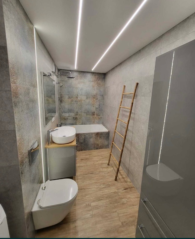 Nowoczesna łazienka z szarą mozaiką, podwieszaną toaletą, umywalką nablatową na szafce z drewnianym blatem, bambusową drabinką i podświetleniem LED na suficie i ścianie.