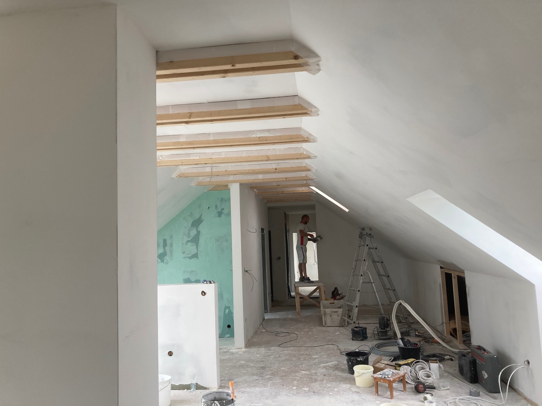 Poddasze w trakcie prac wykończeniowych: szpachlowanie ścian, drewniane belki zabezpieczone folią, drabina, narzędzia malarskie i elektryczne, widoczny pracownik na podwyższeniu.