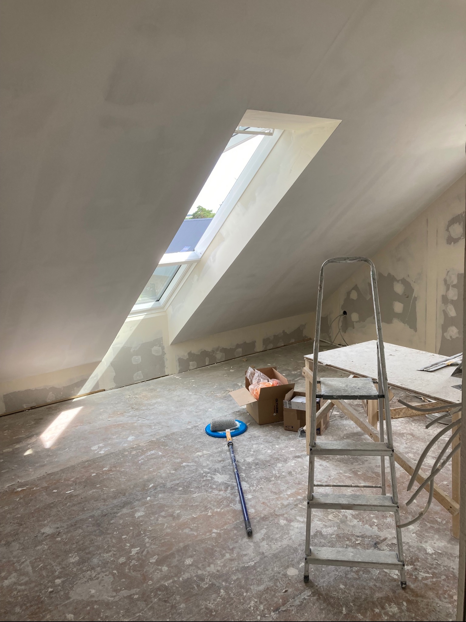 Poddasze w trakcie prac wykończeniowych: wyszpachlowane ściany, okno dachowe, drabina aluminiowa, wałek malarski na podłodze, widoczne ślady szpachli na podłodze, przygotowane kartony i stół roboczy.