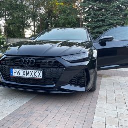 ISS - Studio Auto Detailingu Kevin Kaczmarek - Czarny samochód Audi RS6 zaparkowany na kostce brukowej, z otwartymi drzwiami kierowcy, widoczne logo Audi na grillu i tablica rejestracyjna P6 XMXKX, w tle zieleń drzew i krzewów.
