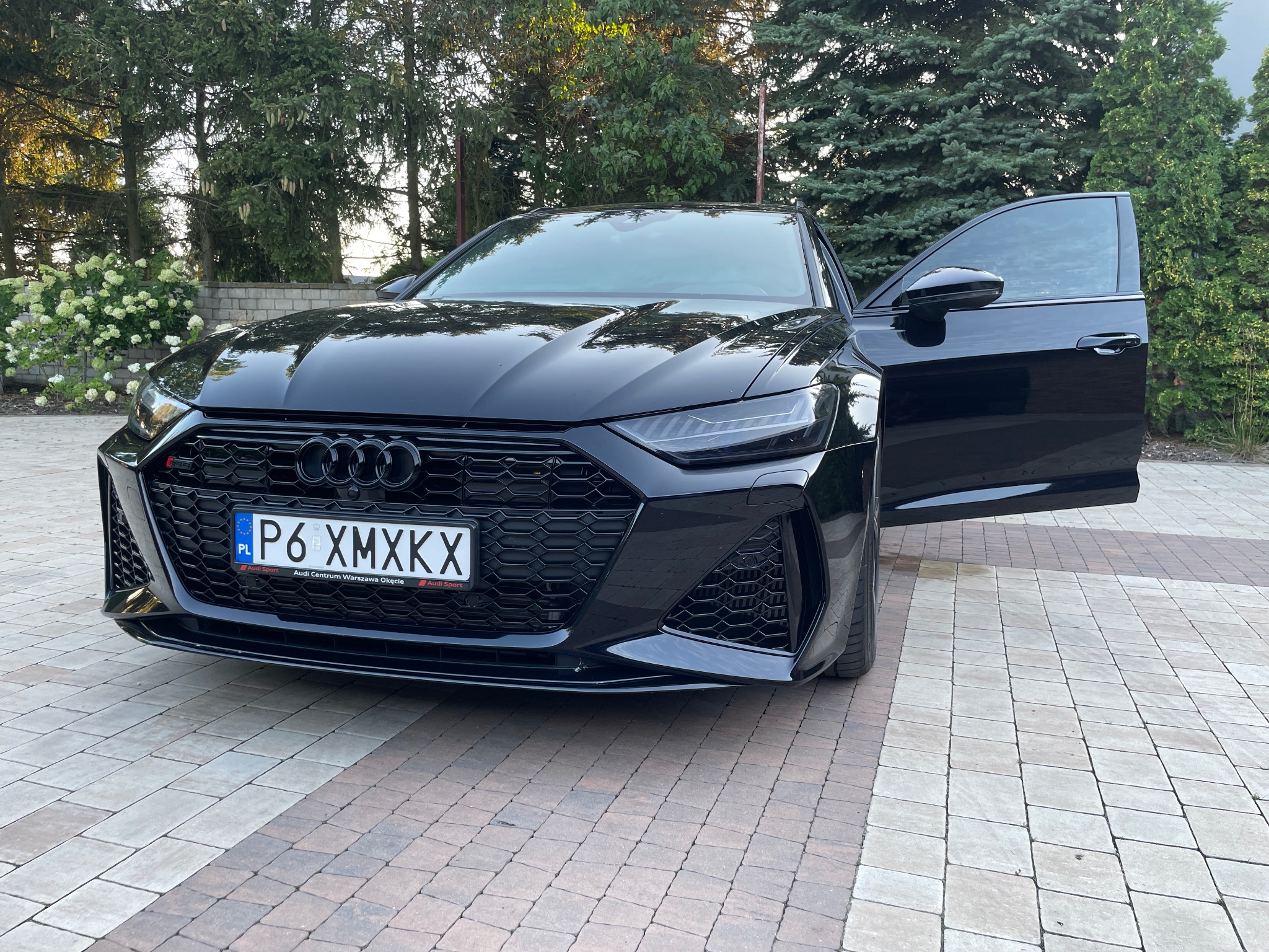 Czarny samochód Audi RS6 zaparkowany na kostce brukowej, z otwartymi drzwiami kierowcy, widoczne logo Audi na grillu i tablica rejestracyjna P6 XMXKX, w tle zieleń drzew i krzewów.