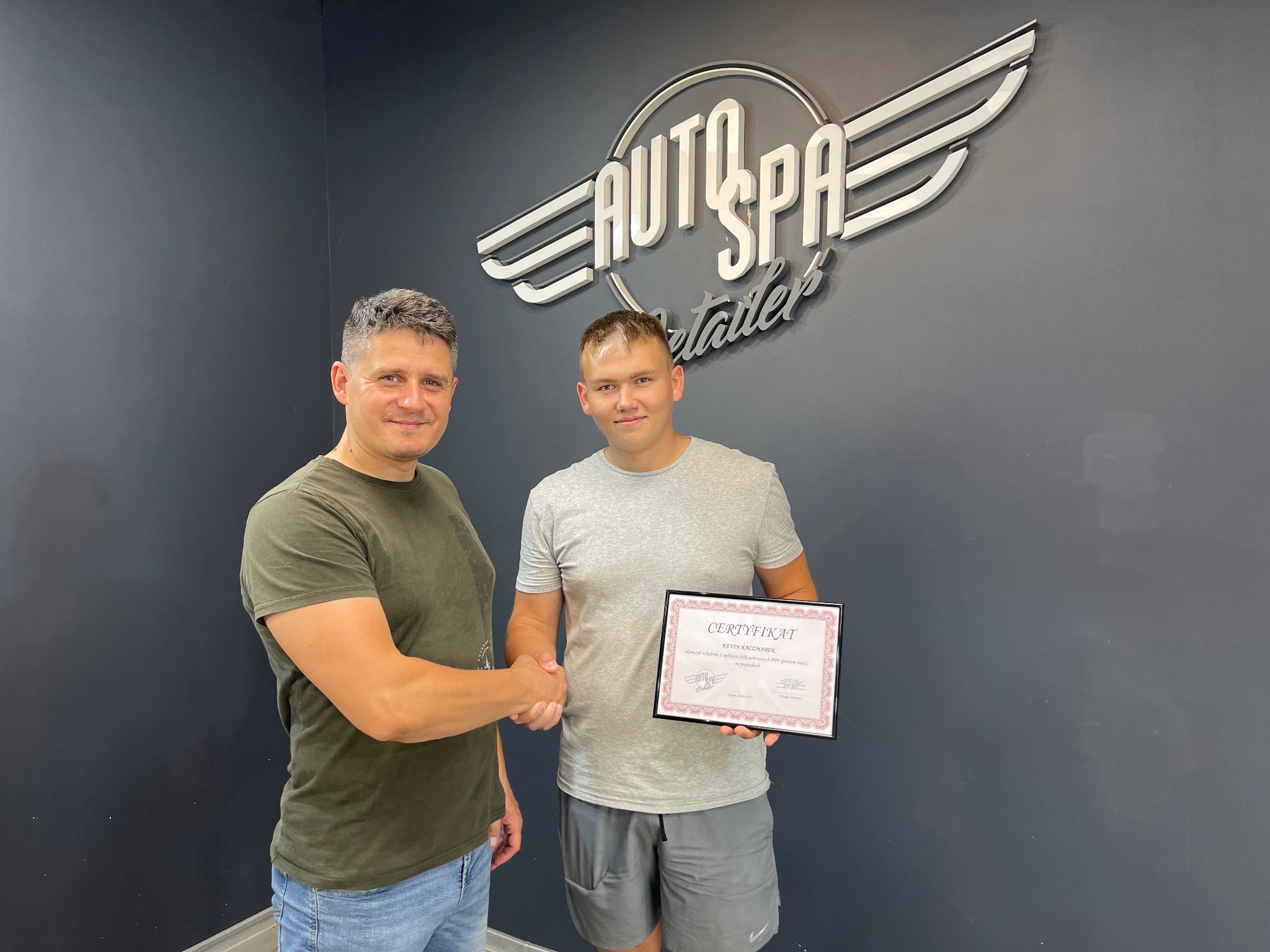 Dwaj mężczyźni w uścisku dłoni, jeden trzyma certyfikat ukończenia szkolenia Auto Spa Detailer na tle logo firmy.