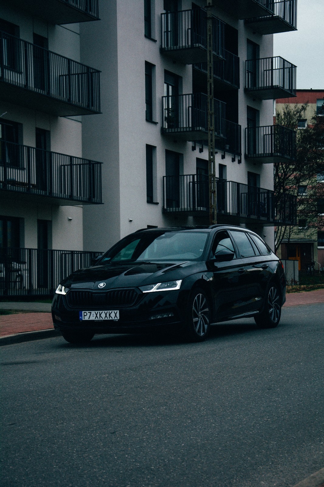Czarny samochód marki Skoda Octavia Combi zaparkowany na ulicy przed nowoczesnym budynkiem mieszkalnym z balkonami.