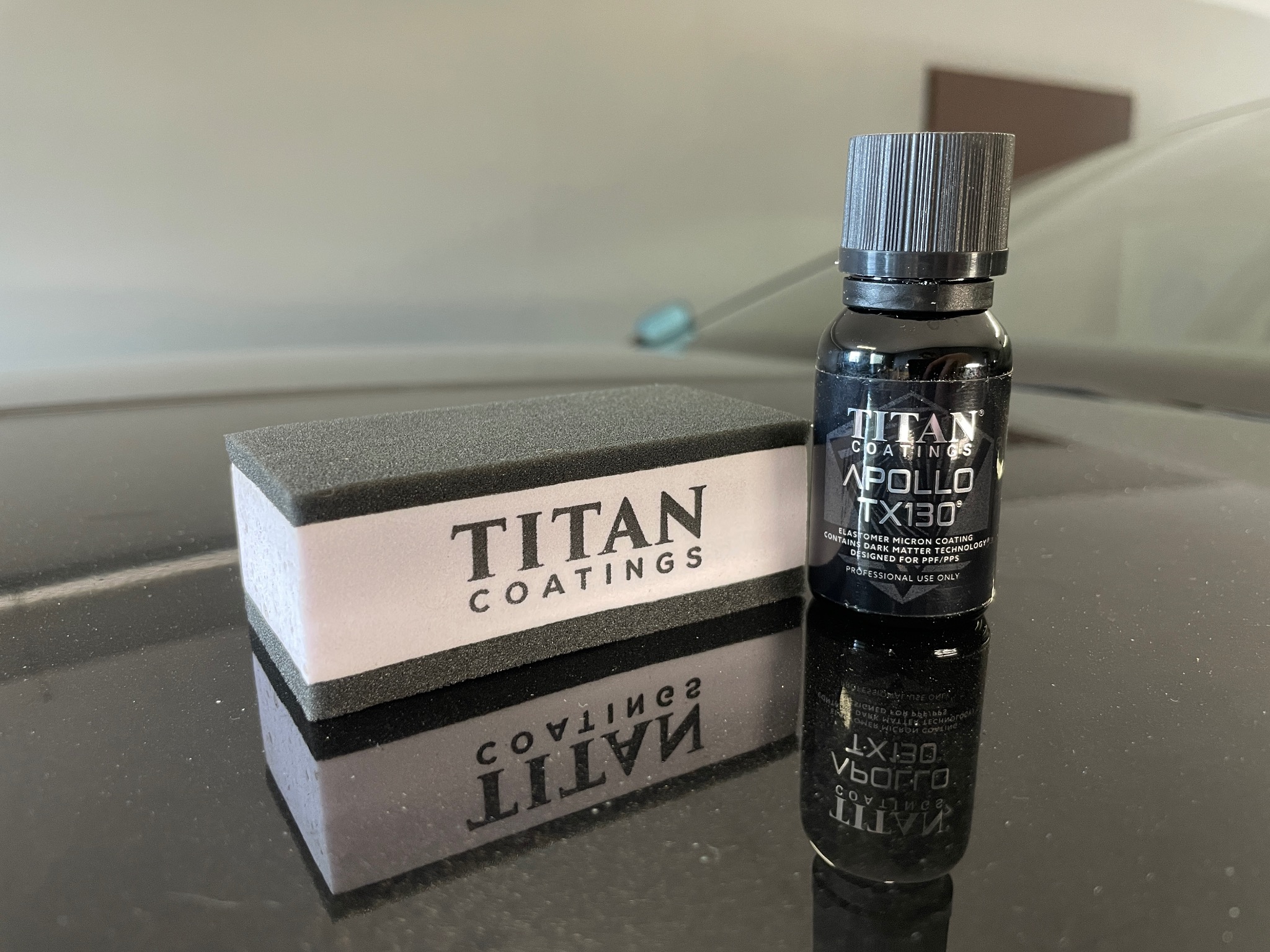 Butelka czarnego preparatu Titan Coatings Apollo TX130 i aplikator z logo Titan Coatings na błyszczącej czarnej powierzchni, odbicie w powierzchni.