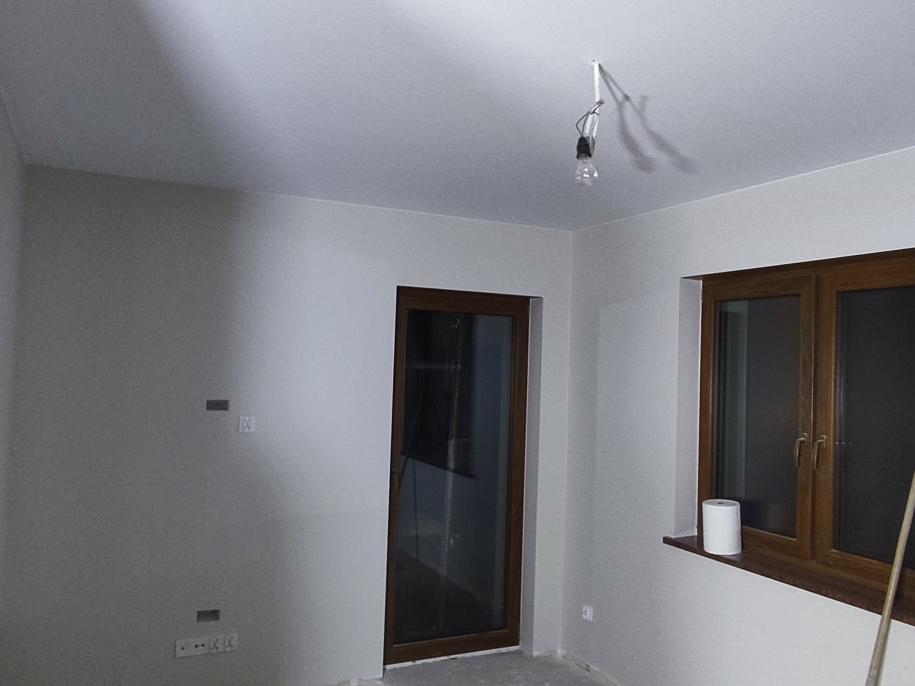 Pokój po szpachlowaniu ścian i sufitu, widoczne gniazdka elektryczne, okno z brązową ramą i wisząca żarówka bez klosza.