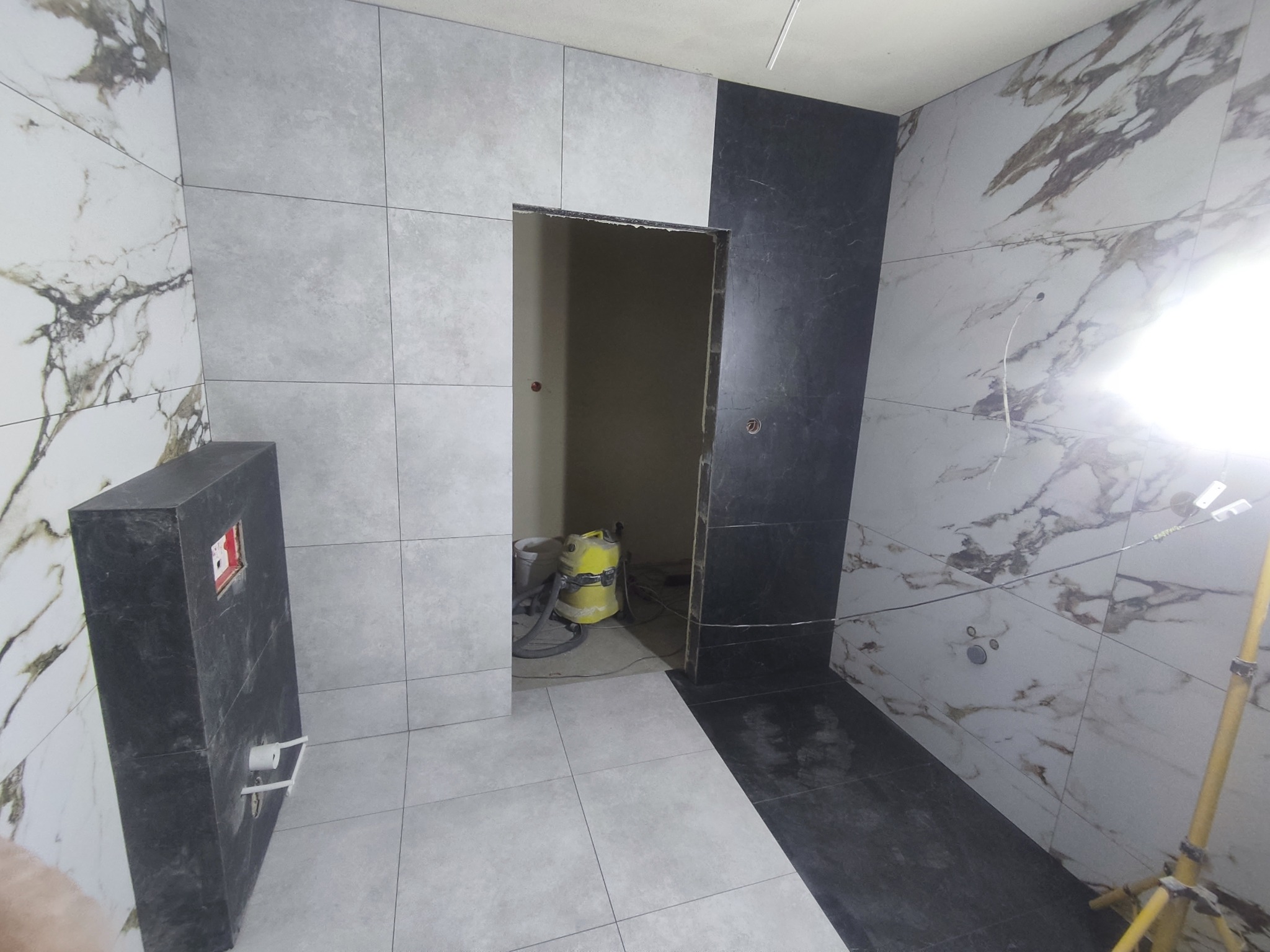 Wykończenie łazienki w trakcie: szare i marmurowe płytki na ścianach i podłodze, widoczny fragment czarnej obudowy WC, odkurzacz przemysłowy w przejściu do innego pomieszczenia, lampa budowlana