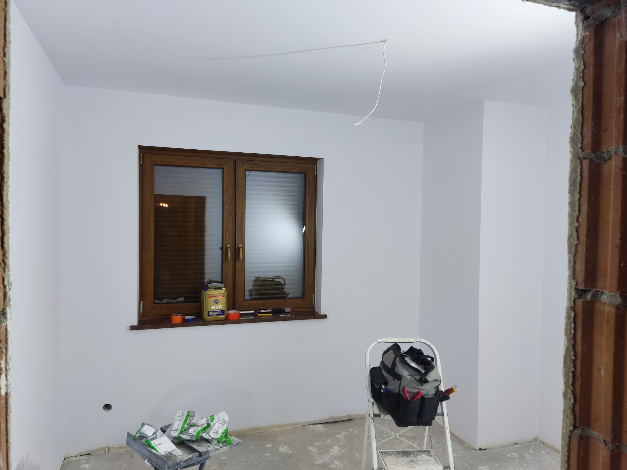 Świeżo pomalowane na biało wnętrze pokoju z brązowym oknem, na parapecie puszka farby INKP, drabina z torbą narzędziową i resztki materiałów na podłodze.