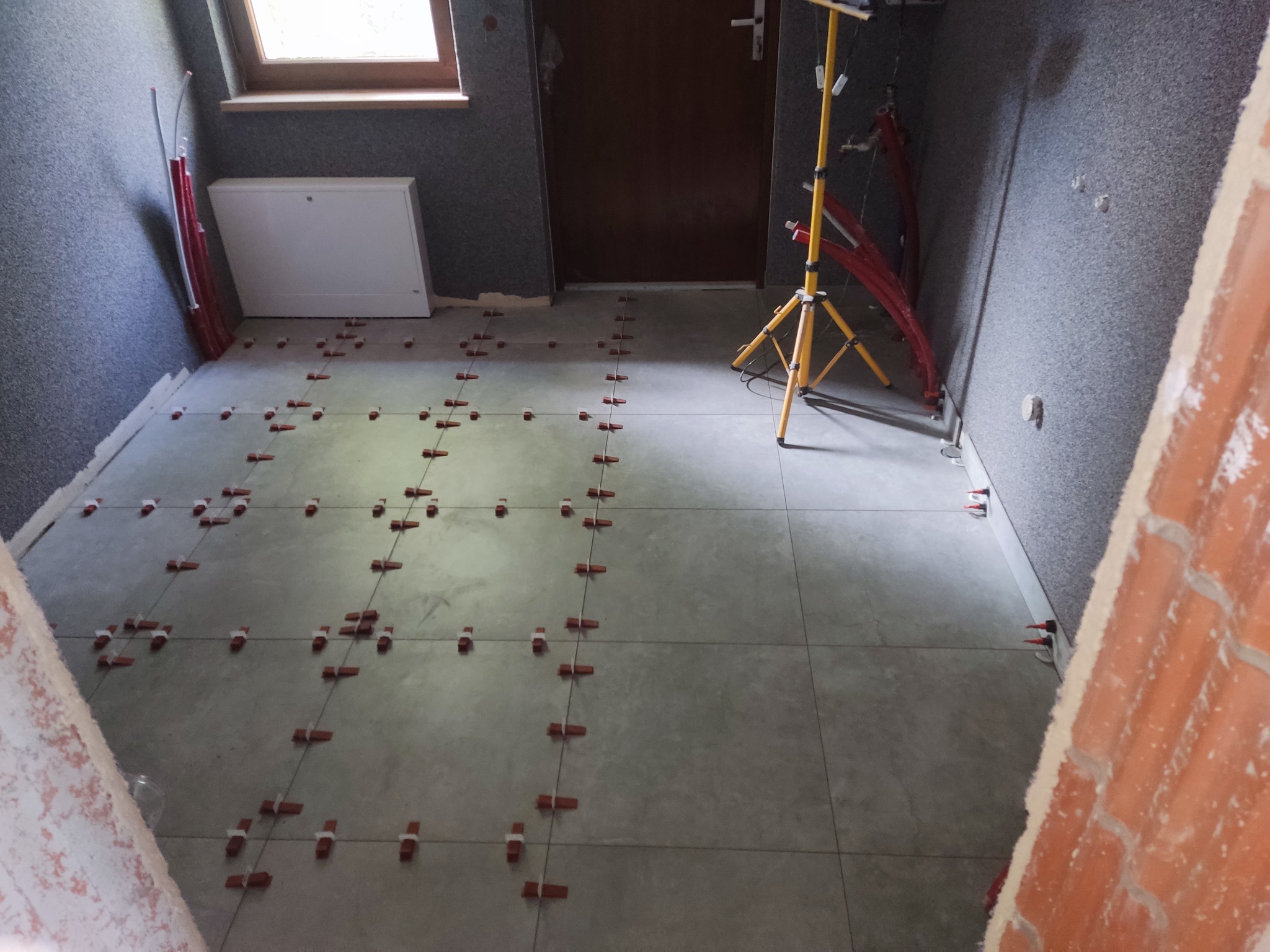 Układanie szarych płytek podłogowych z użyciem czerwonych krzyżyków dystansowych, widoczna lampa budowlana i fragment ściany z szorstką fakturą.