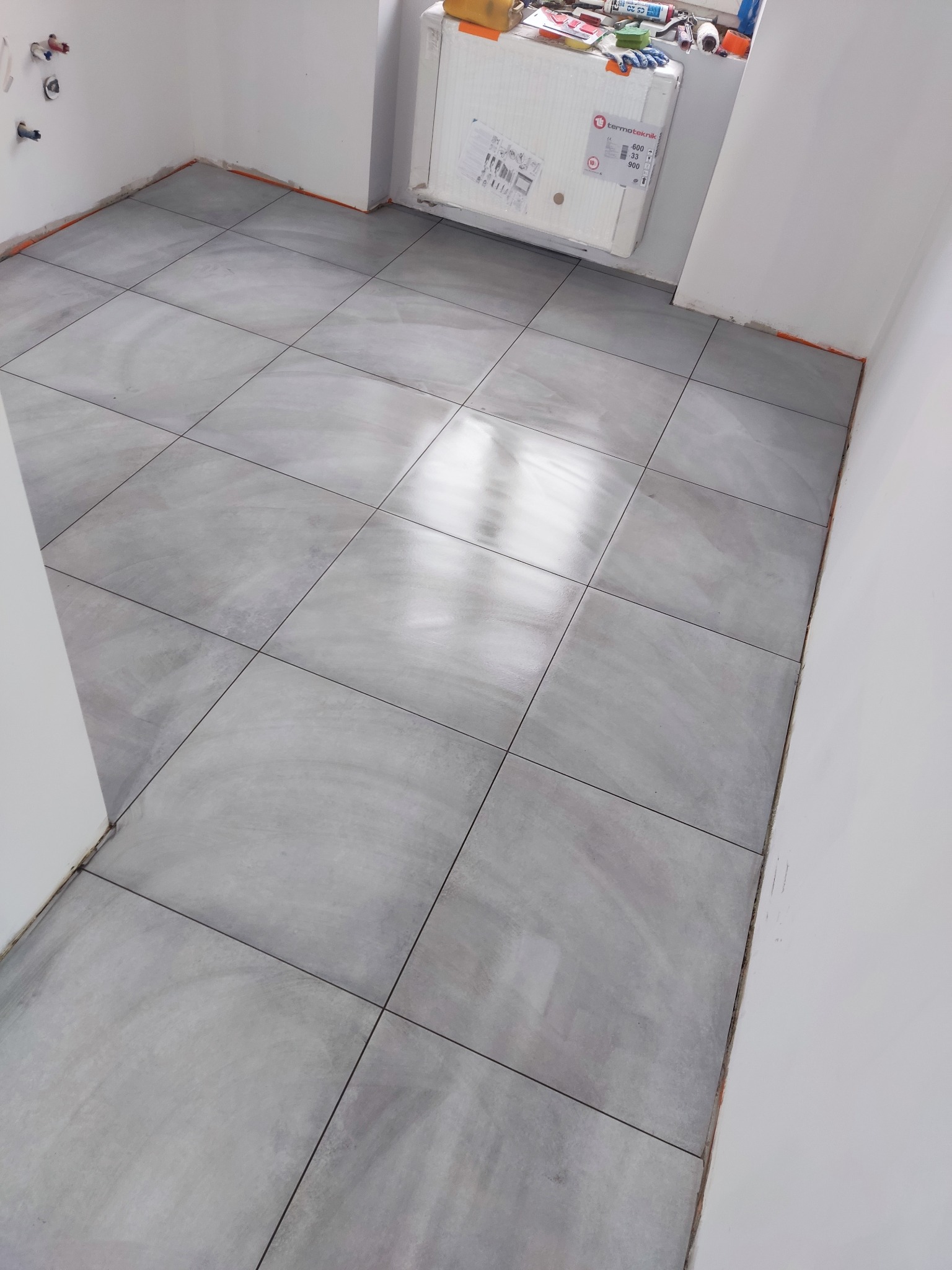 Ułożone szare płytki podłogowe w łazience, widoczne niedokończone ściany i grzejnik z narzędziami na parapecie okna.